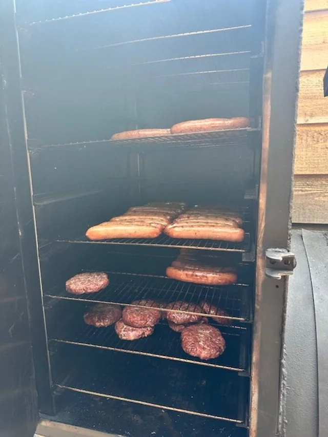 Meat patties and sausages cooking on grill racks inside a smoker or barbecue grill.