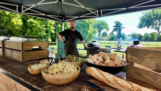 Man standing outdoors under a canopy with various food items on a wooden table, including pasta, bread, and bowls of salad, with a grill and open landscape in the background.