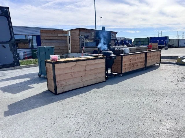 Outdoor food stand with wooden counters, a grill emitting smoke, and multiple containers for food or supplies. Storage sheds and shipping containers in the background.