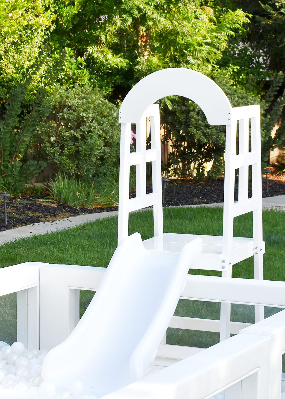 A white platform plastic slide leading into a ball pit, with a white wooden climbing frame and an arch in a backyard garden with green bushes, trees, and a lawn.