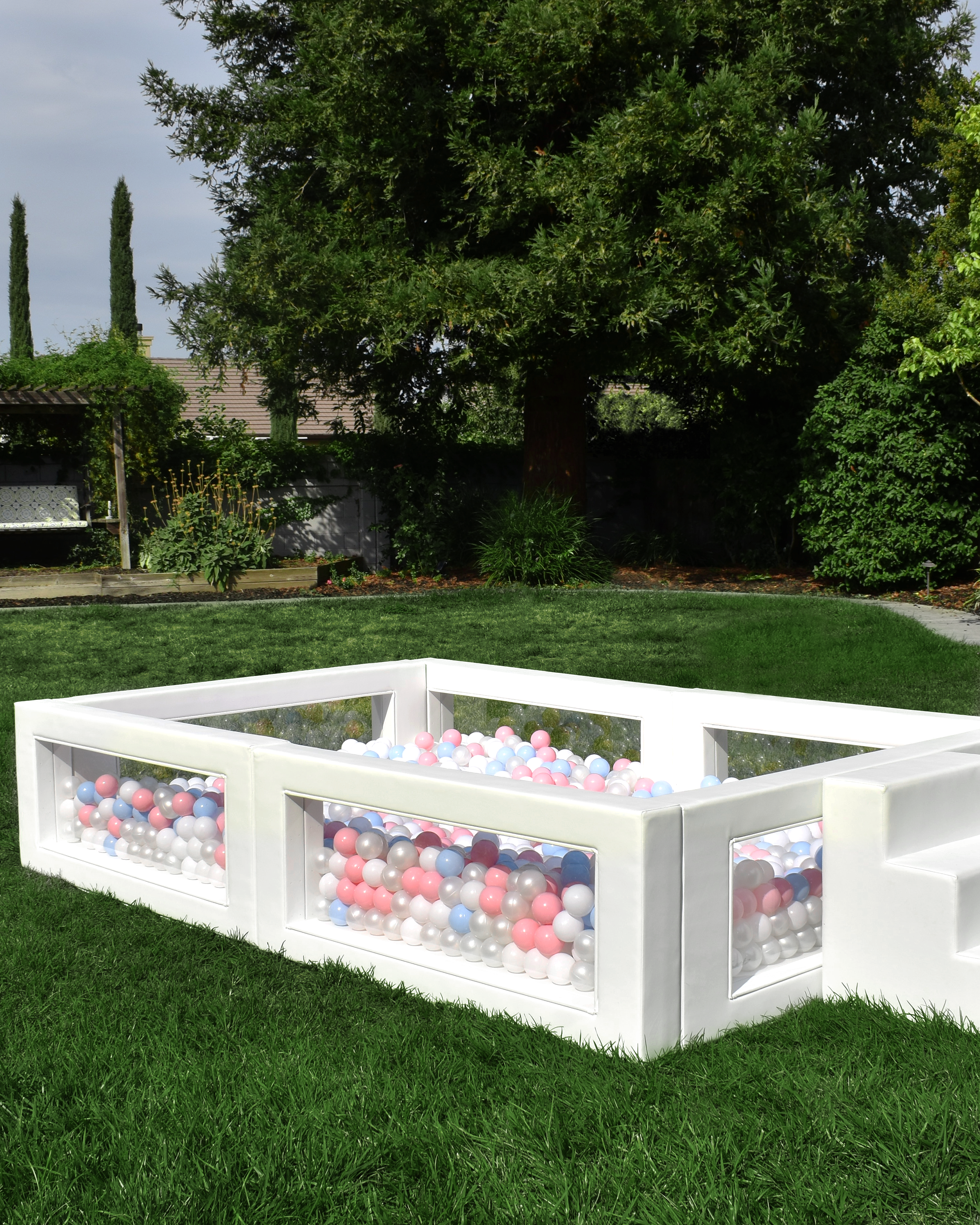 A backyard with green grass, trees, and bushes. In the center, there is a white, rectangular ball pit filled with pink, white, blue, and gray plastic balls, surrounded by steps.