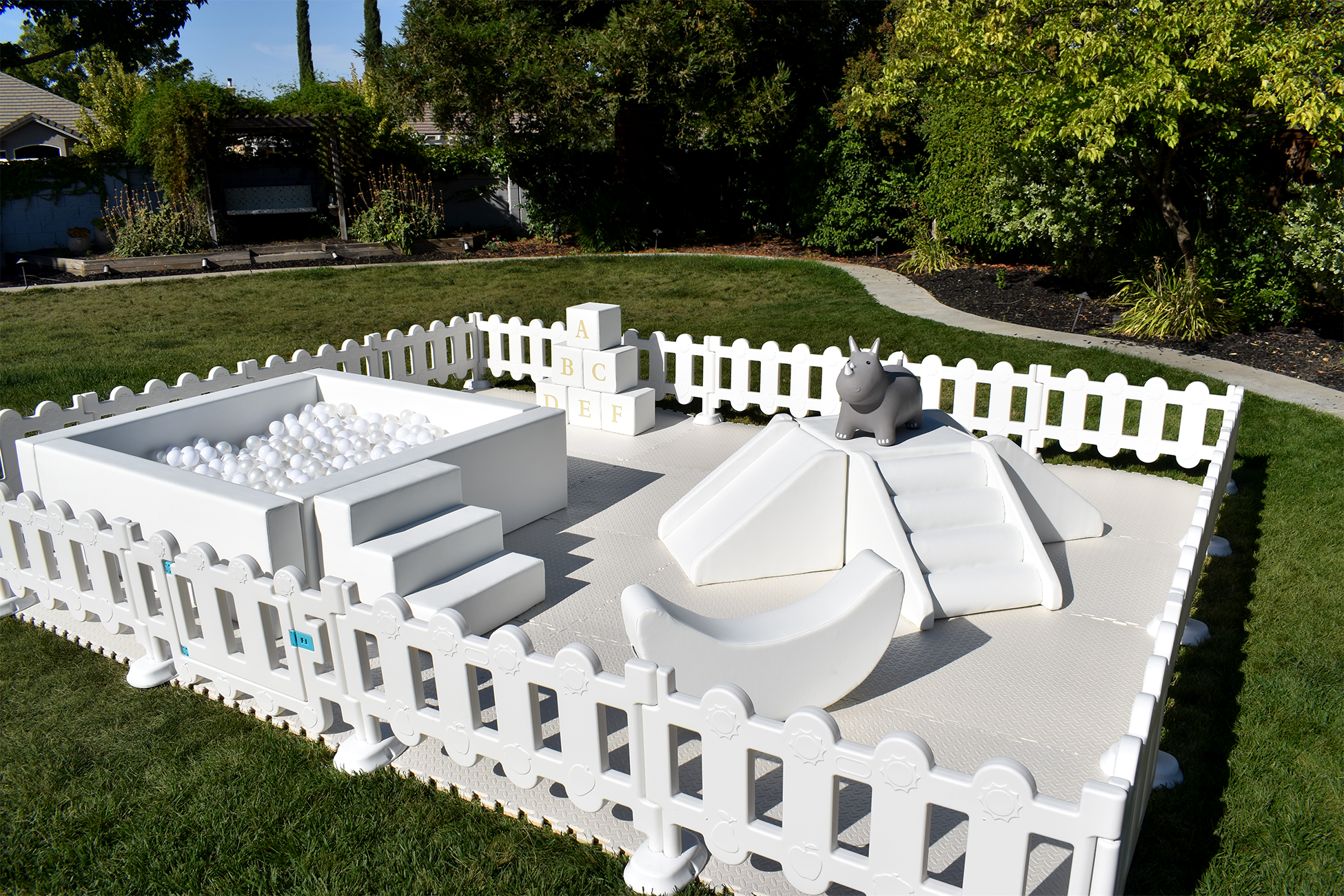 Outdoor soft play area for children with a ball pit, stairs, slide, and animal toy, surrounded by a white picket fence on a grassy yard.