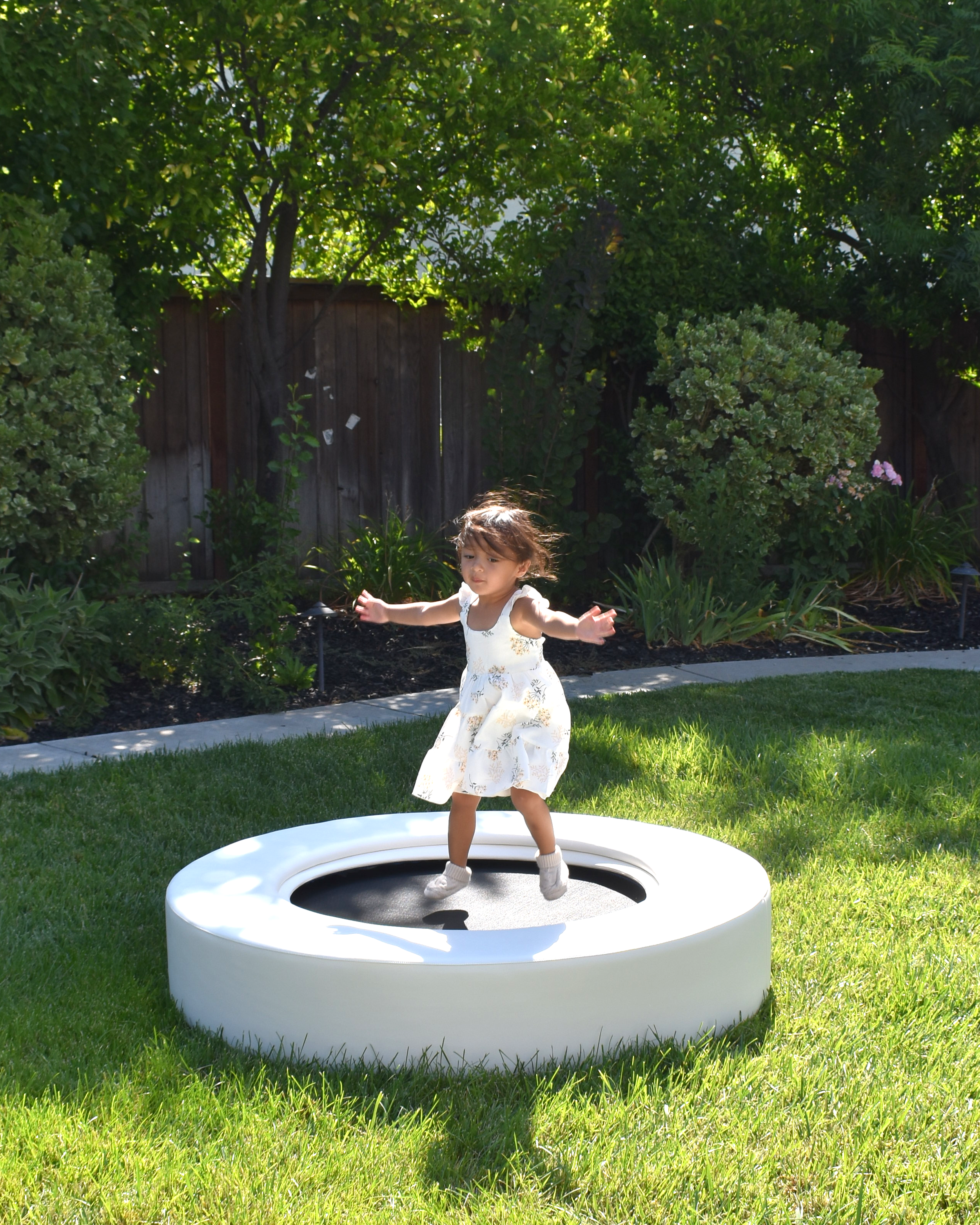 A young girl in a white dress bouncing on a small trampoline in a backyard with green grass, trees, bushes, and a wooden fence.