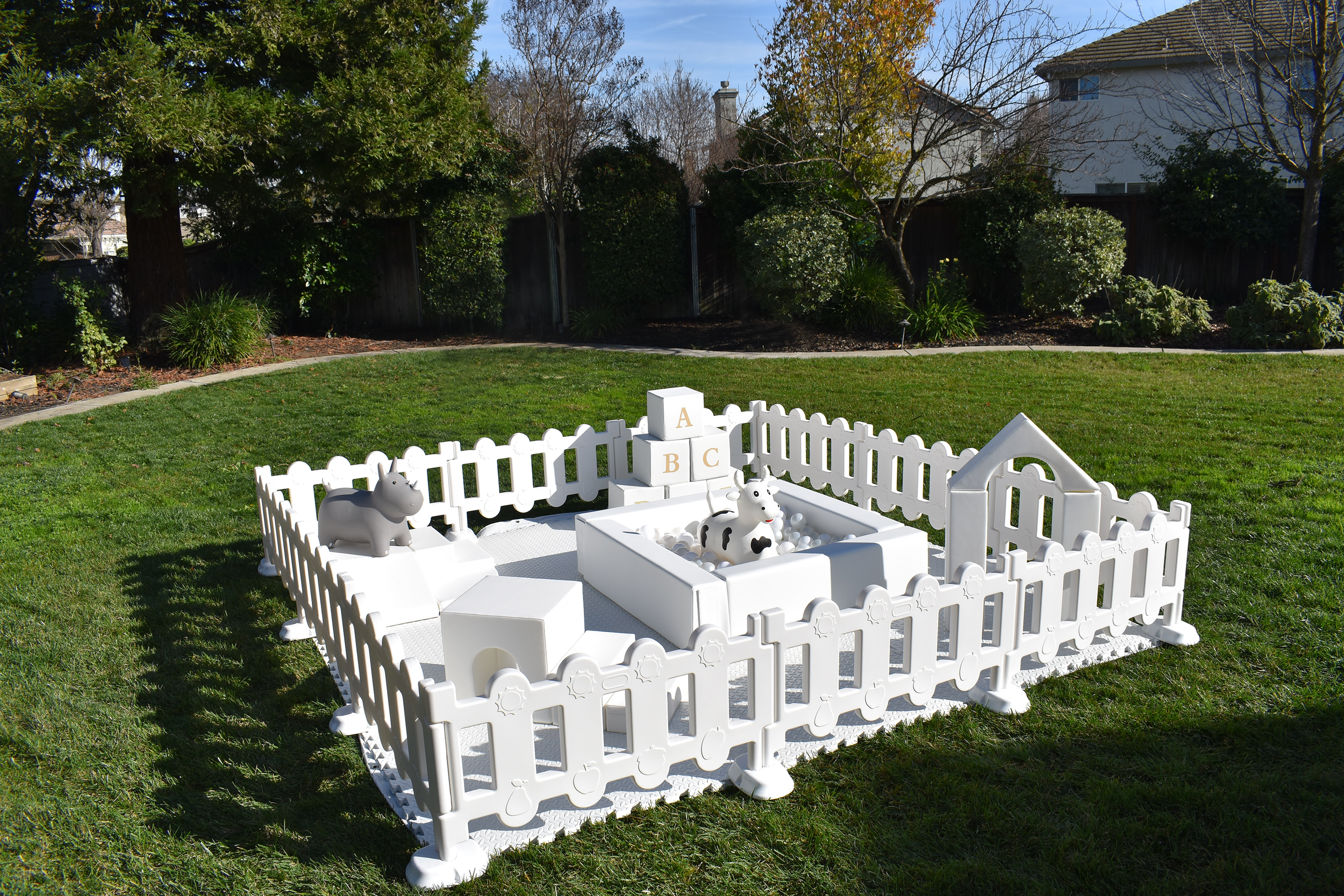 Outdoor play area with a white plastic fence, cow toys, alphabet blocks, and a small house structure on a grass lawn with trees and bushes in the background.