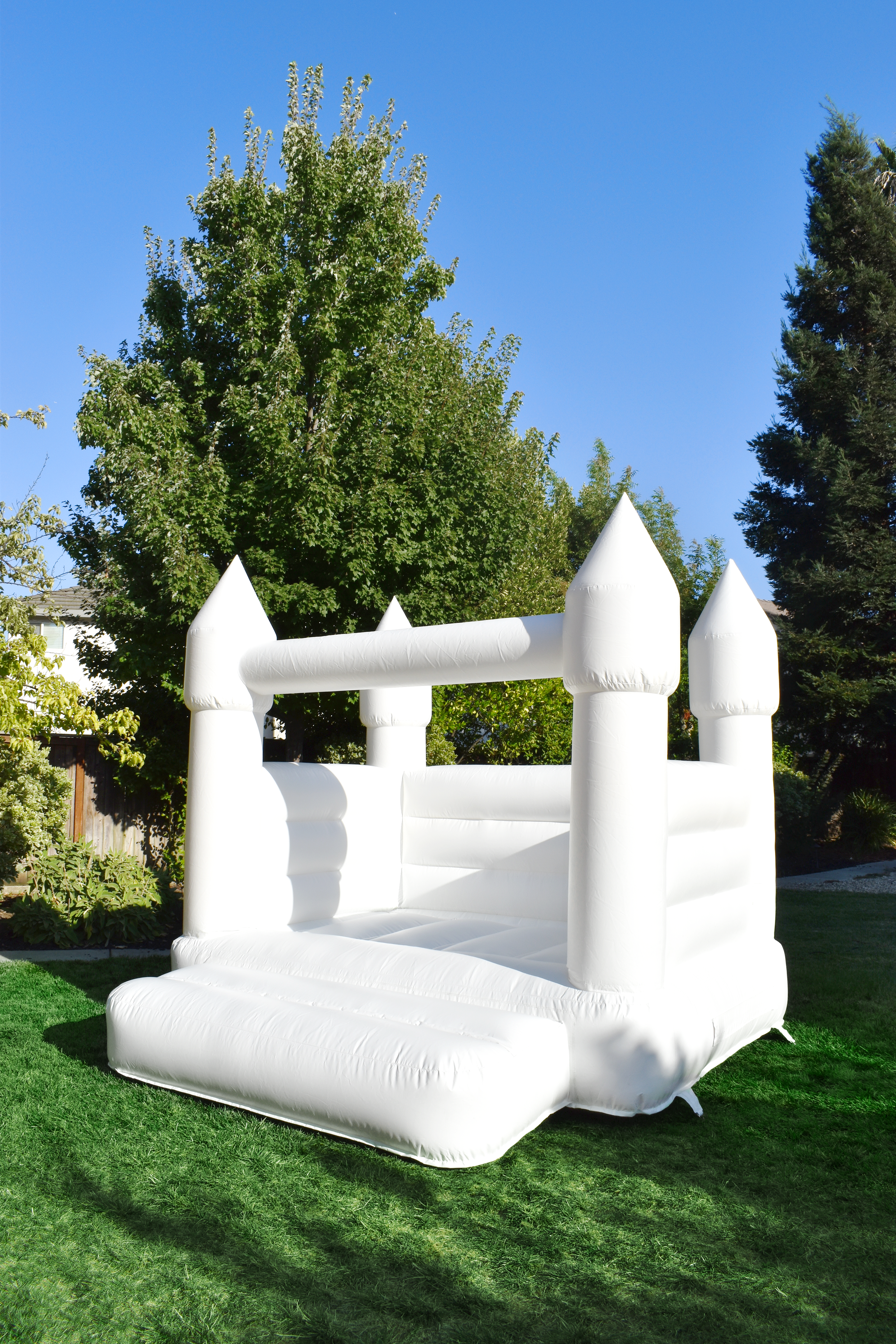 A white inflatable bounce house designed like a castle with four towers, set up on a green lawn in a backyard, with trees and a clear blue sky in the background.