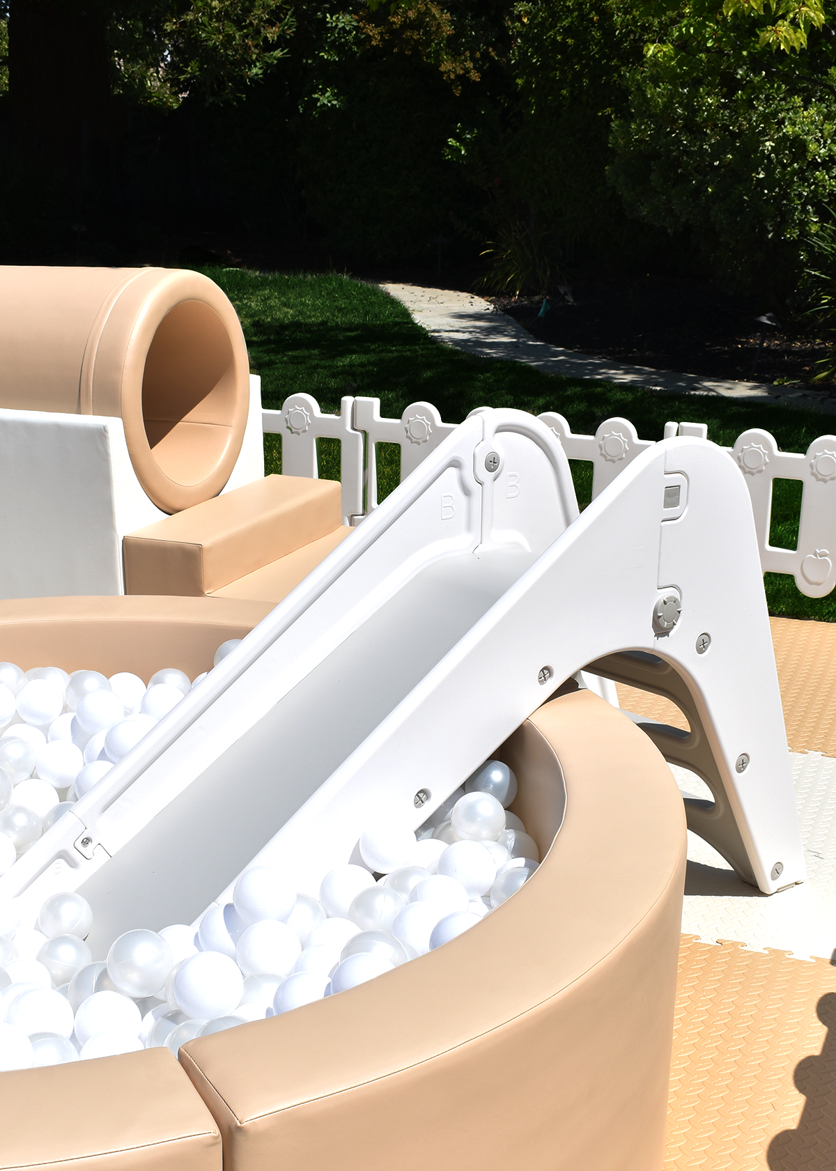 Outdoor soft play set with a beige slide filled with white plastic balls, surrounded by a white fence and green grass with trees in the background.