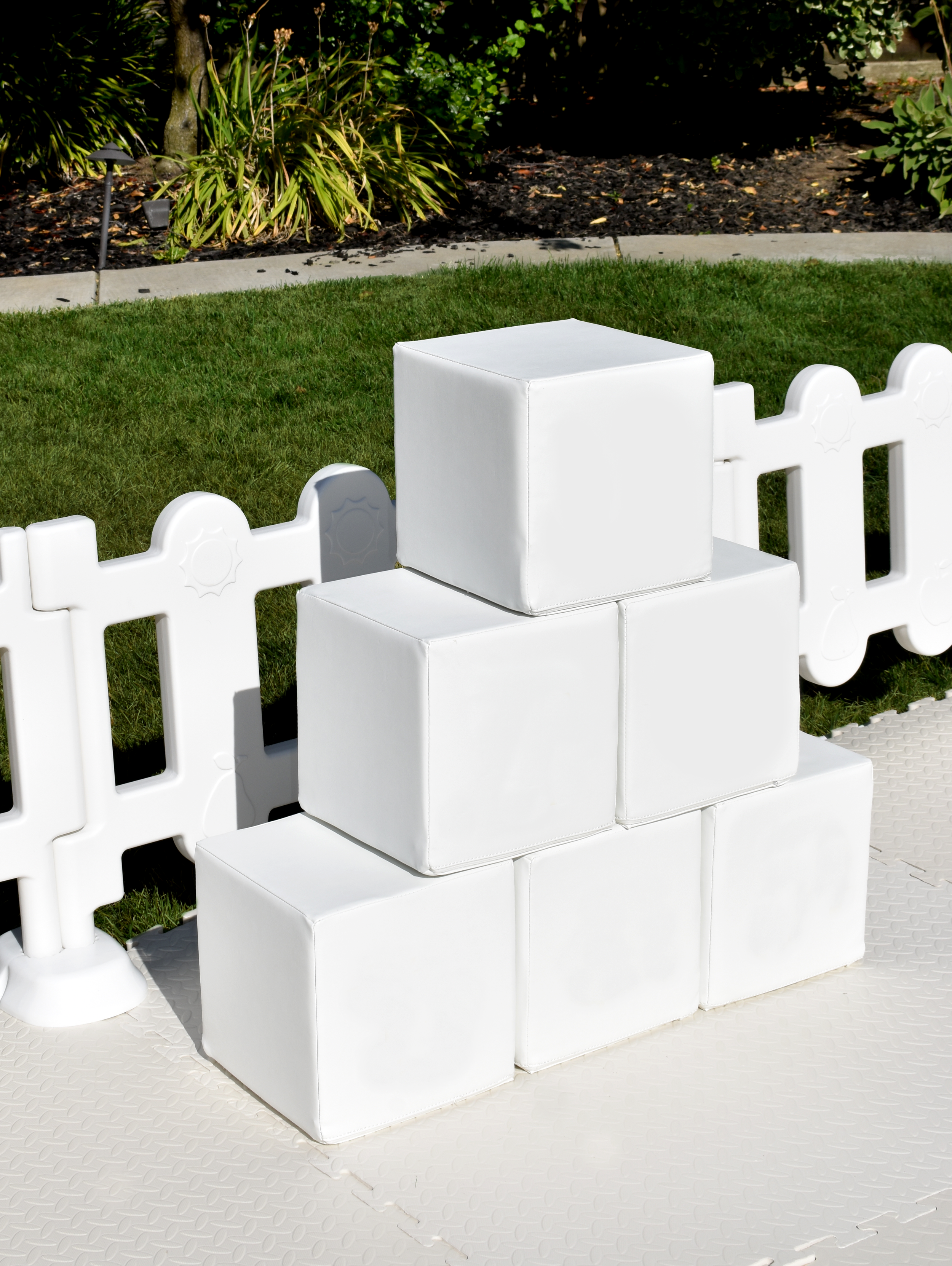 Stacked white foam blocks arranged in a pyramid shape outside on a white deck, with a white picket fence and green bushes in the background.