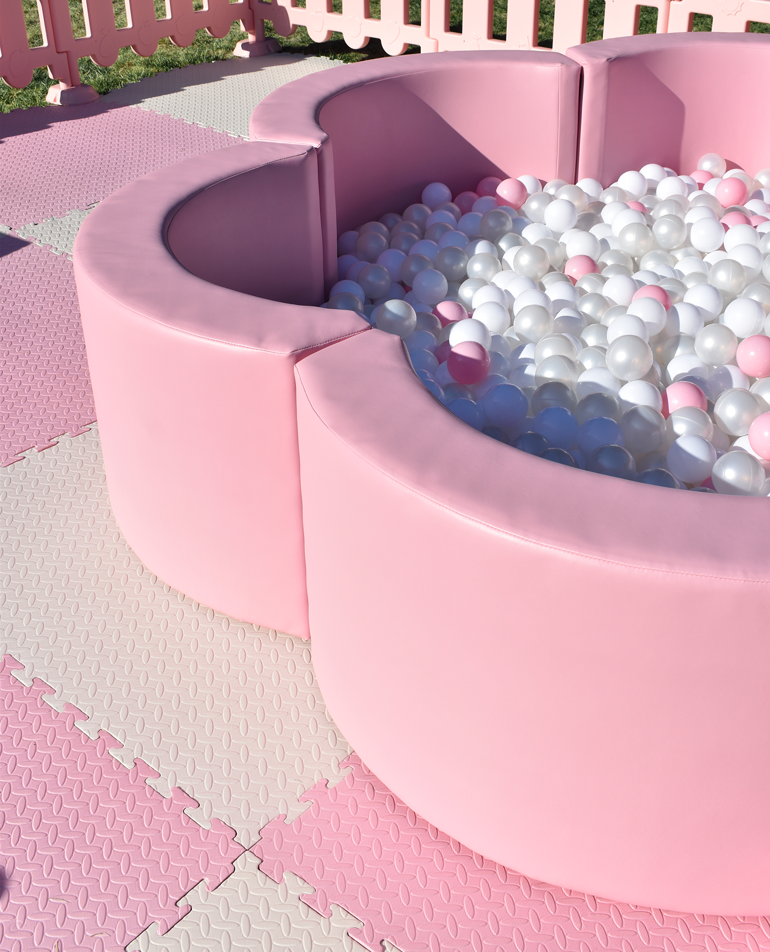 A pink ball pit filled with white and pink plastic balls, surrounded by pink cushioned walls and pink interlocking foam floor mats.