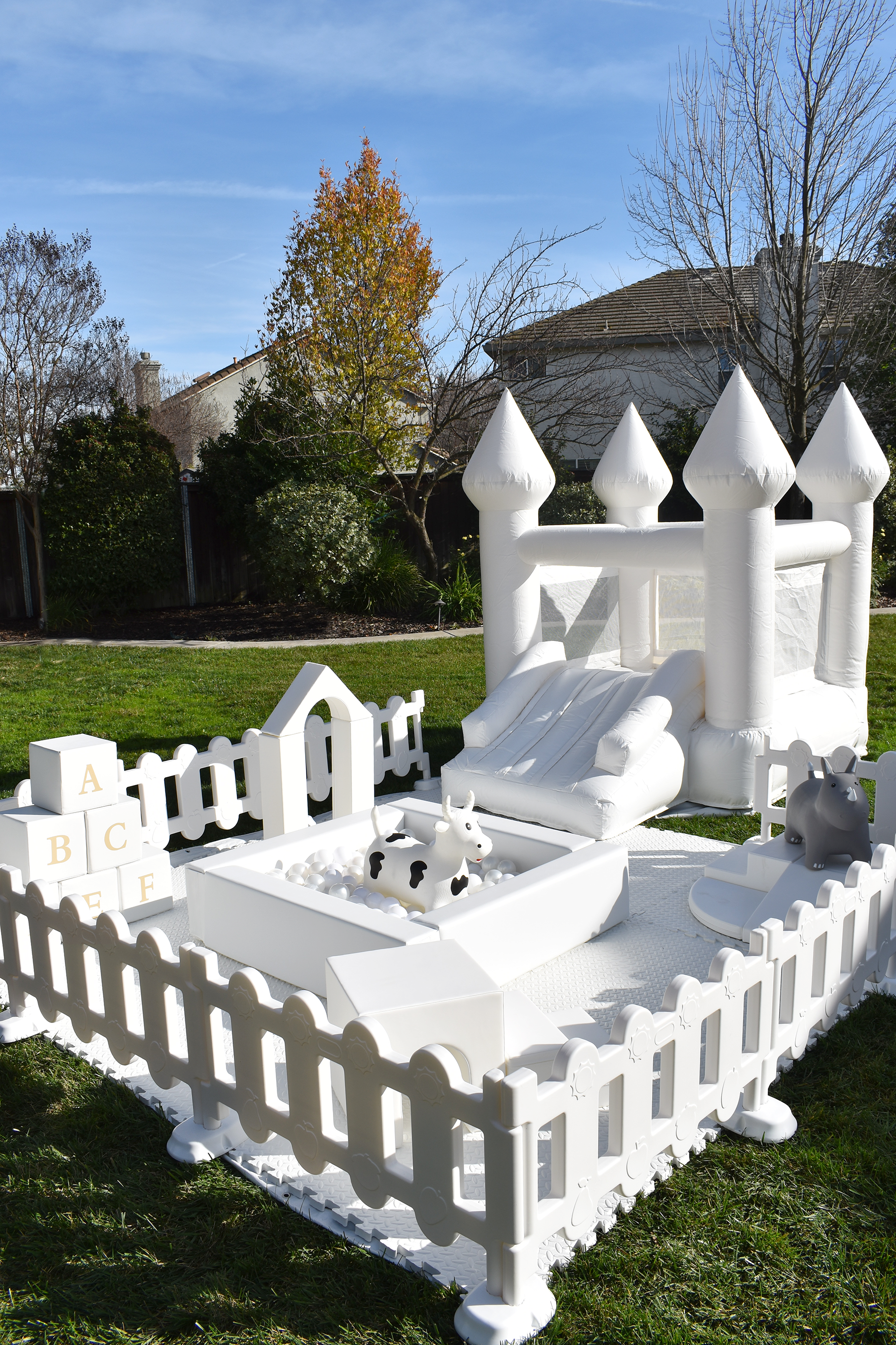 White inflatable bouncy castle with slide in backyard, surrounded by white picket fence with foam blocks, cow, and hippopotamus toys, under blue sky and trees.