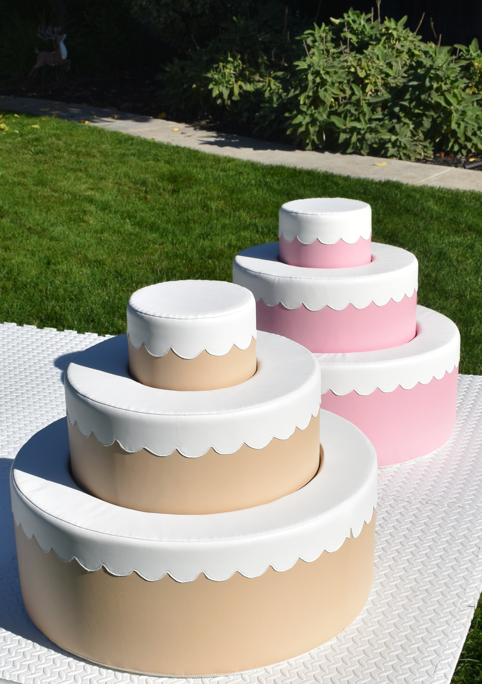 A three-tiered cake decoration in pastel colors, with a beige bottom tier, a pink middle tier, and a white top tier, each topped with a white scalloped border. The cake is on a white textured mat outdoors on grass with plants in the background.