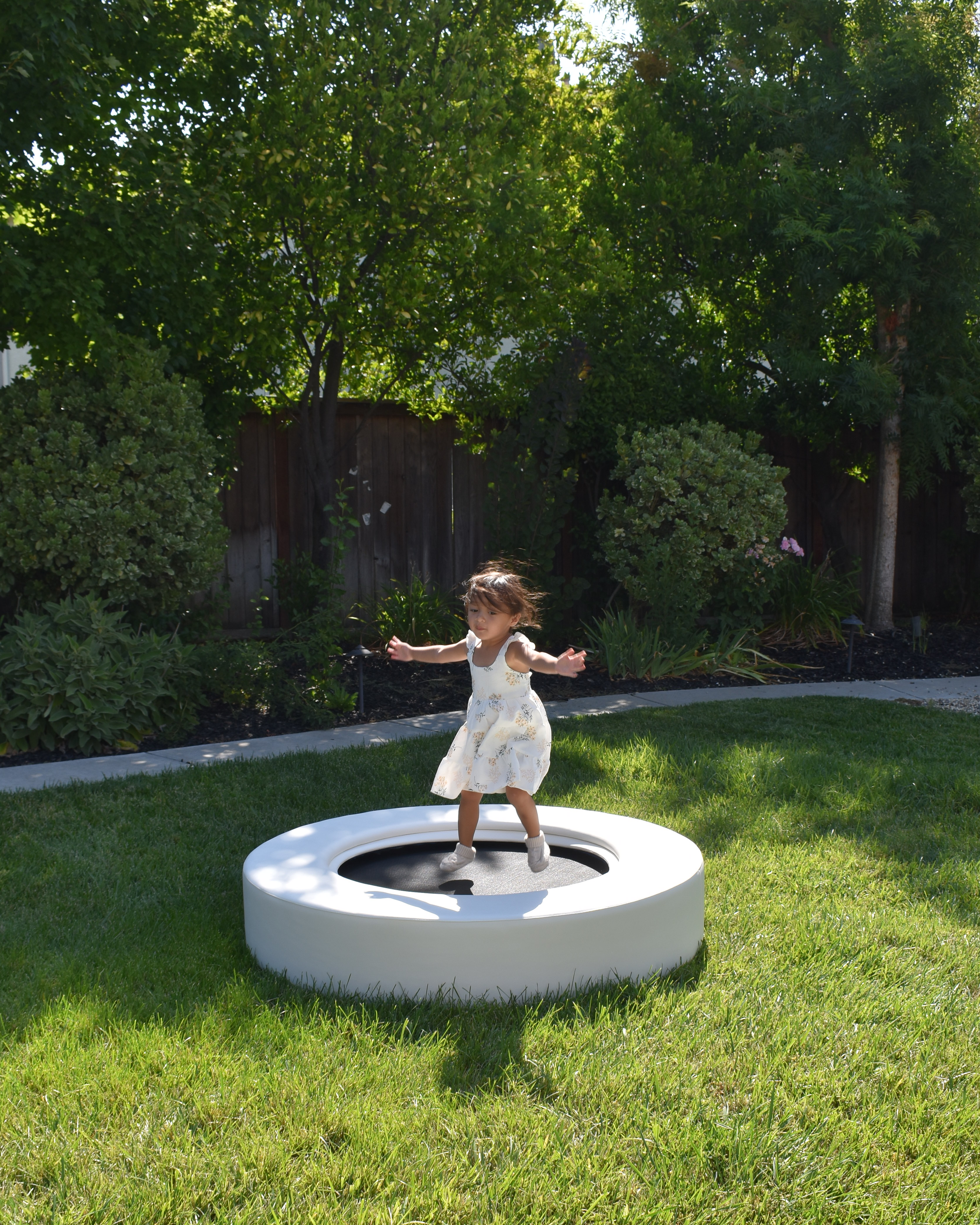 A young girl in a white dress jumping on a small trampoline in a grassy backyard.