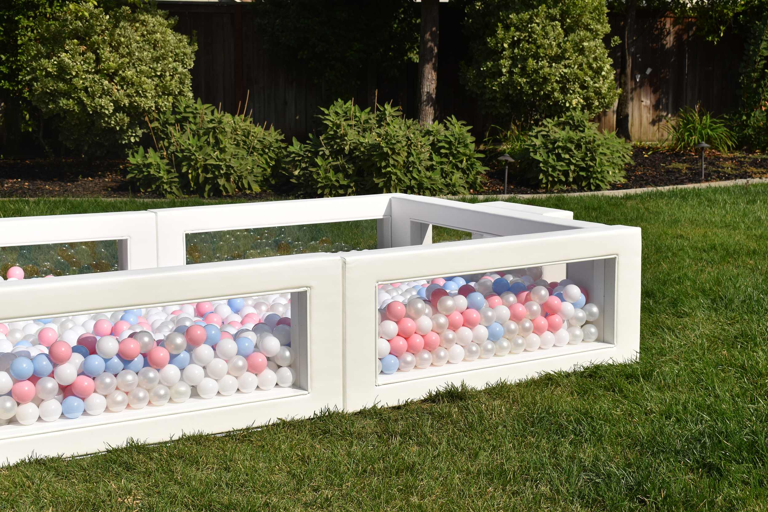 A white outdoor ball pit filled with pink, blue, white, and silver plastic balls, situated on a grassy lawn with bushes and trees in the background.
