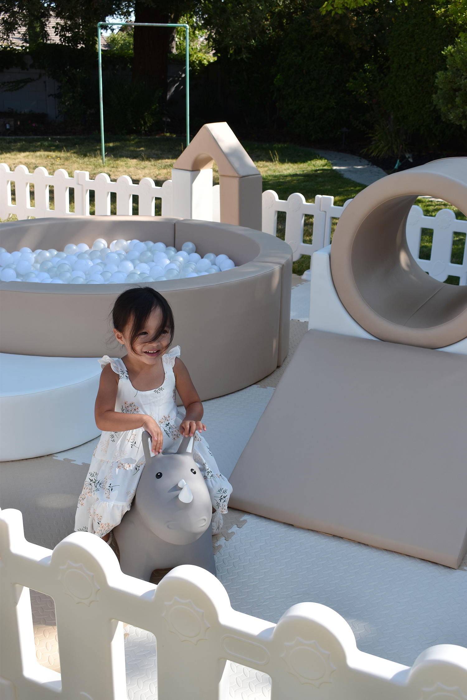 A young girl smiling while playing on a gray toy hippo bounce toy in an outdoor play area with a white fence, ball pit, and playground equipment in the background.