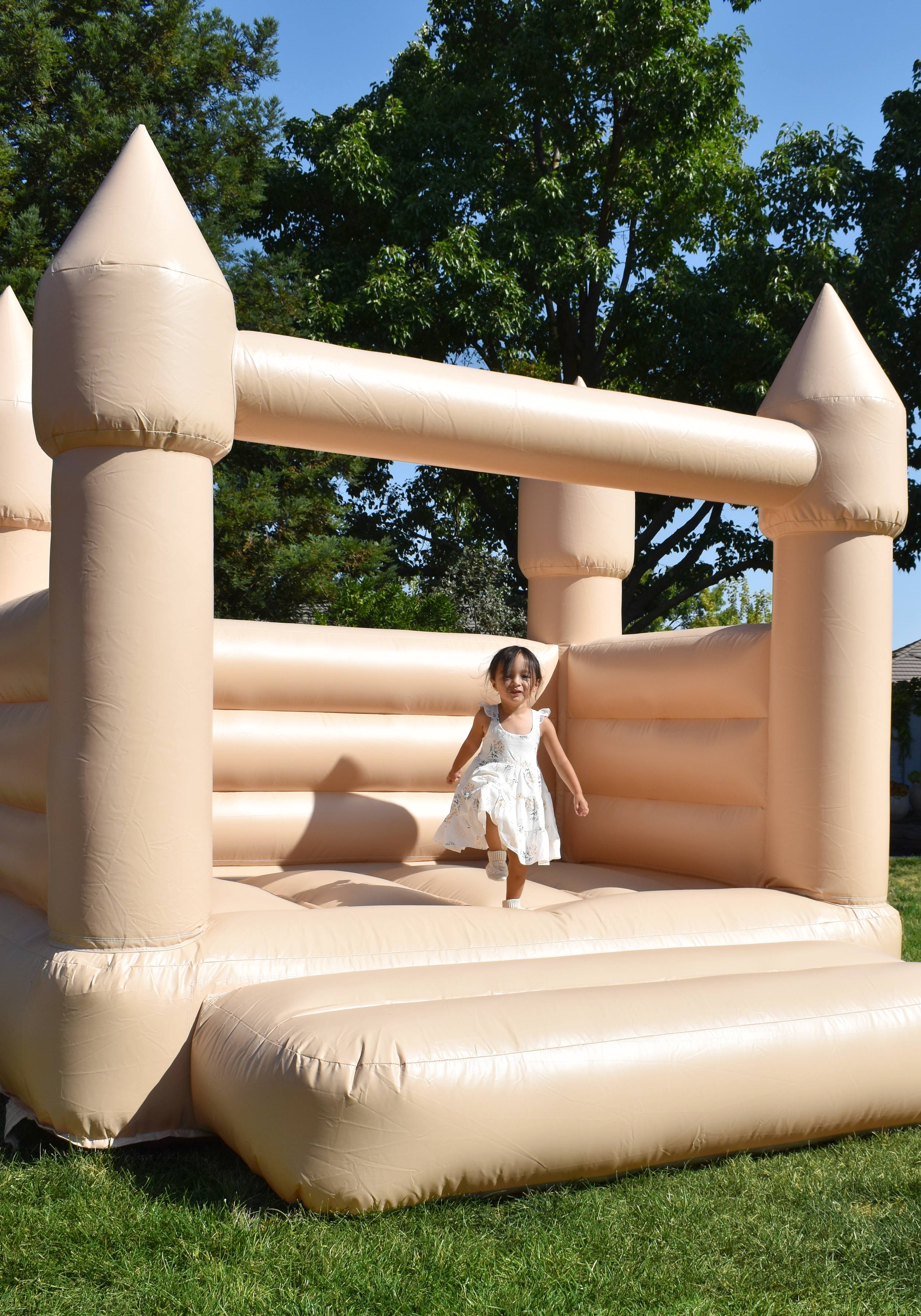 A young girl in a white dress jumping on an inflatable bounce house castle outdoors, surrounded by green trees and a blue sky.
