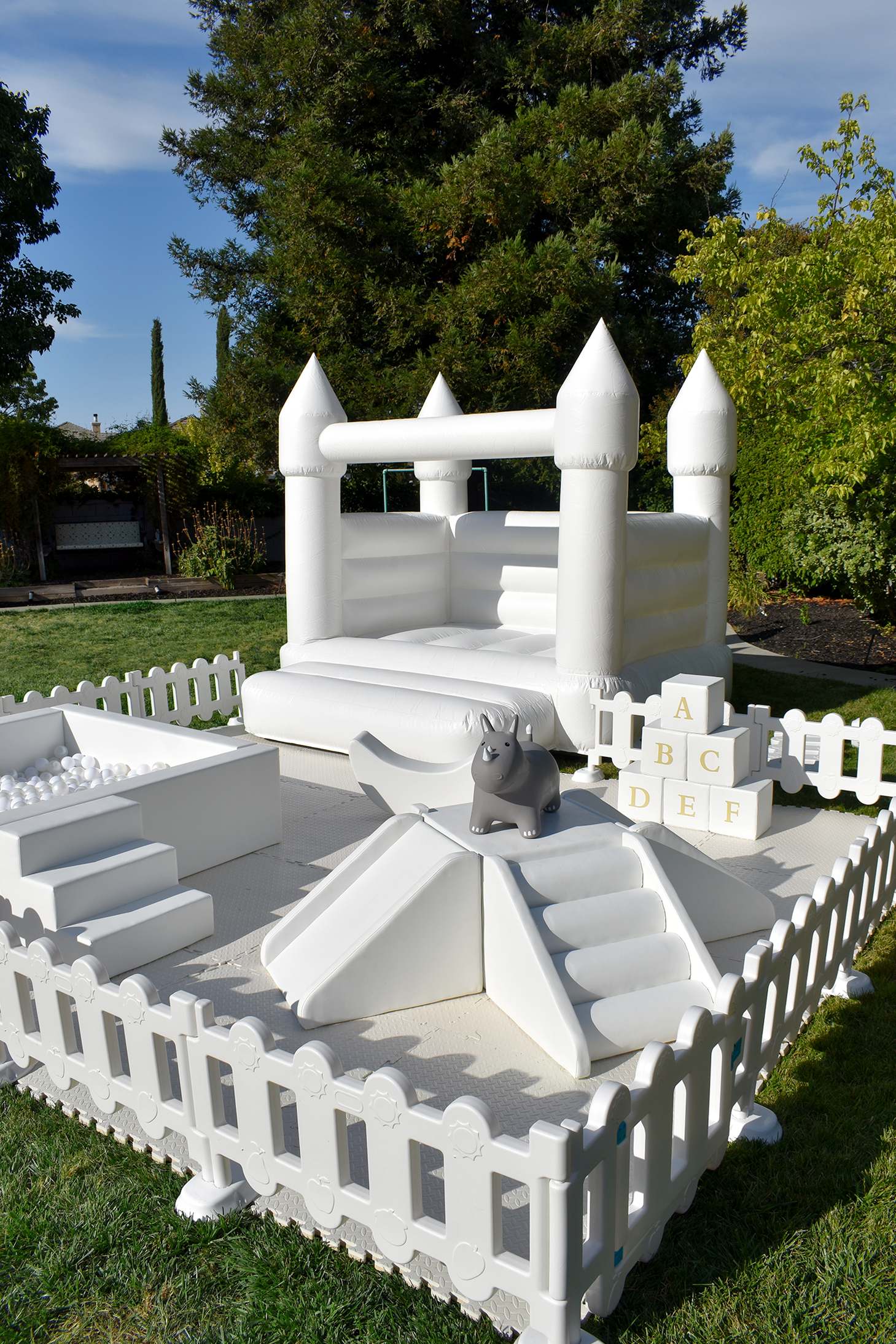 White inflatable bounce house castle with small slide, surrounded by a white picket fence, on a grassy yard. There are toy blocks with letters, a grey toy hippo bouncer, and a ball pit in the area. Big trees and a blue sky are in the background.
