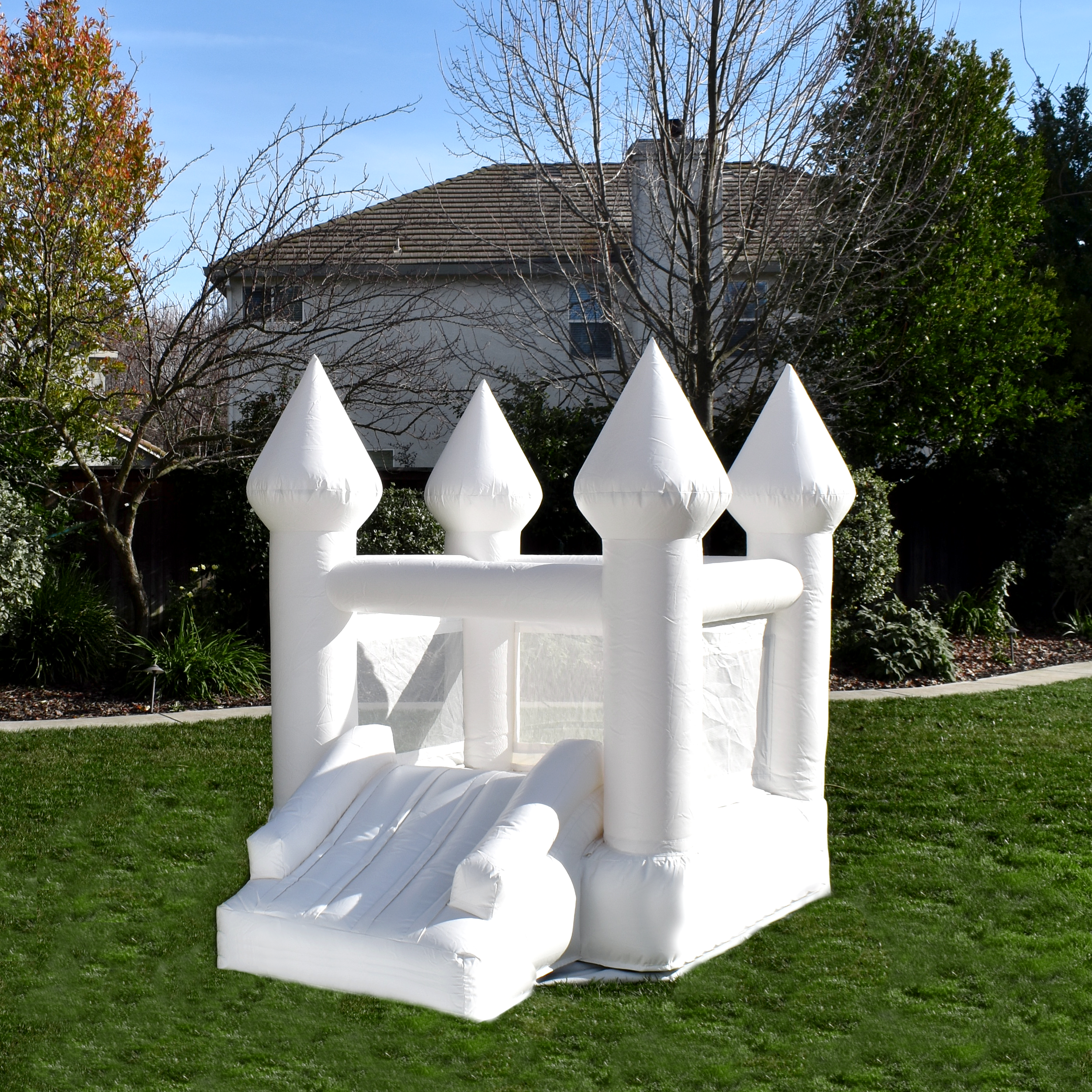 White inflatable castle bounce house in a backyard with grass, trees, and a house in the background.