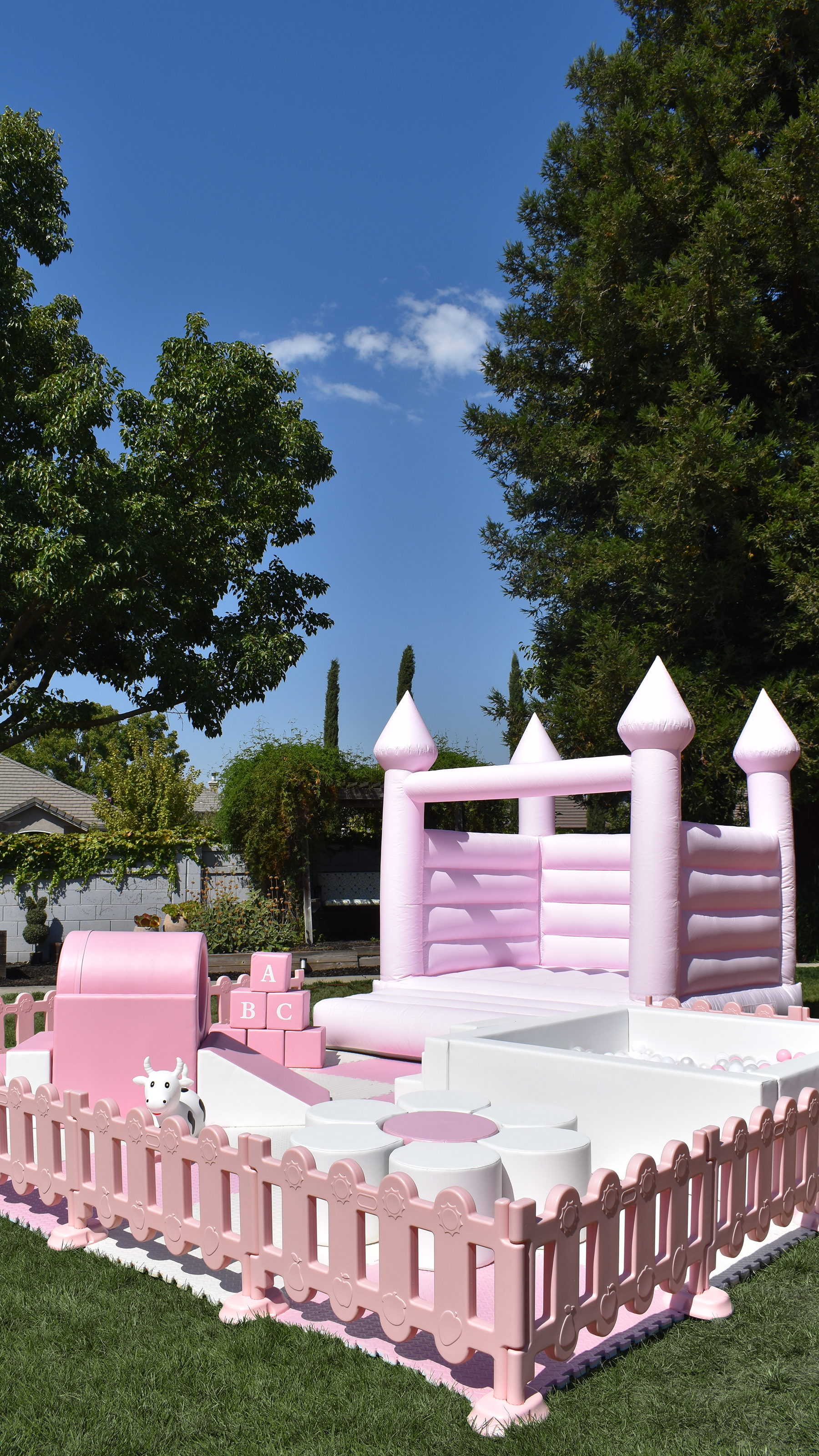 Pink inflatable castle with turrets and a small play area with blocks, a toy cow, and a slide in a backyard surrounded by grass, trees, and a blue sky.