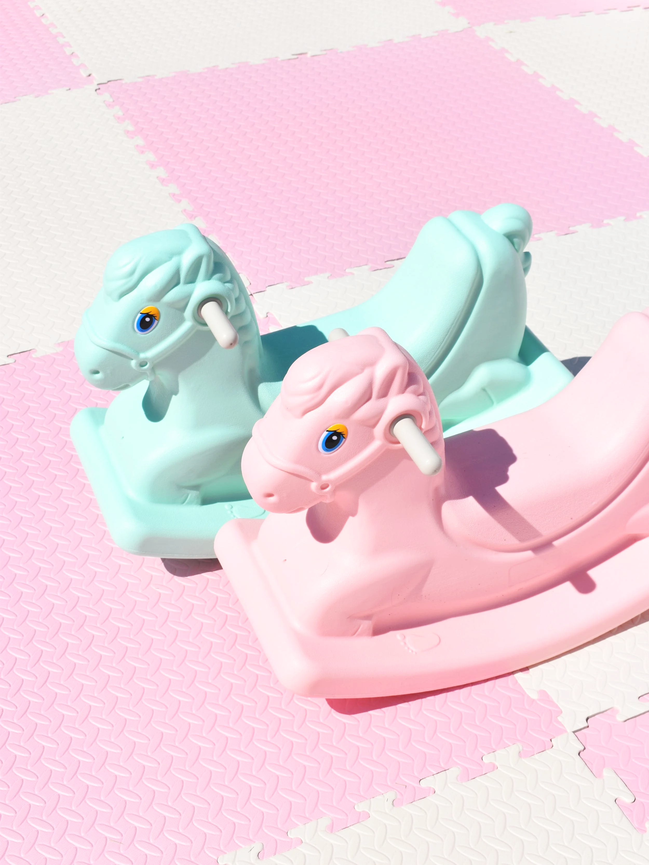Two pastel-colored plastic rocking horses, one mint green and one pink, on a pink and white interlocking foam mat outdoors.
