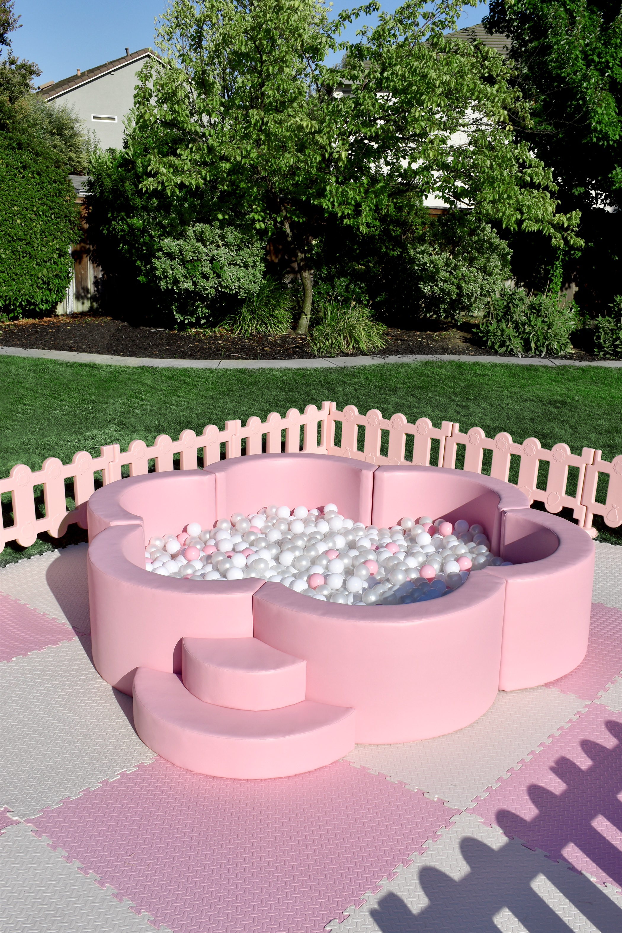 Pink floral ball pit with small steps on a pink and white checkered mat, surrounded by a pink fence, outdoors in a yard with green grass, trees, and houses in the background.