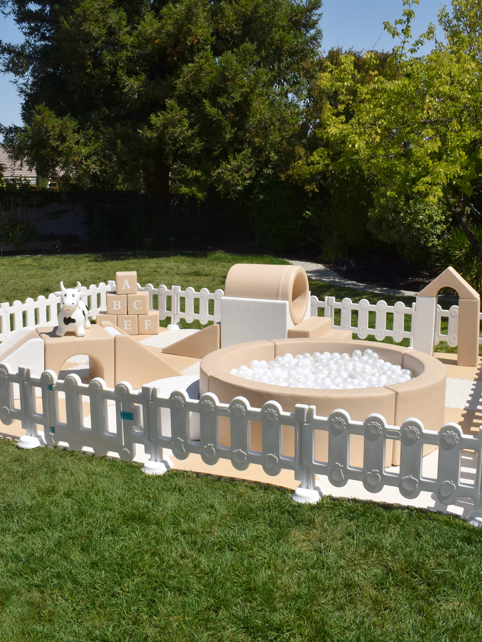 A children's outdoor soft play area with a white picket fence, featuring a ball pit with white balls, a slide, a toy cow, and ABC blocks on grass with trees in the background.