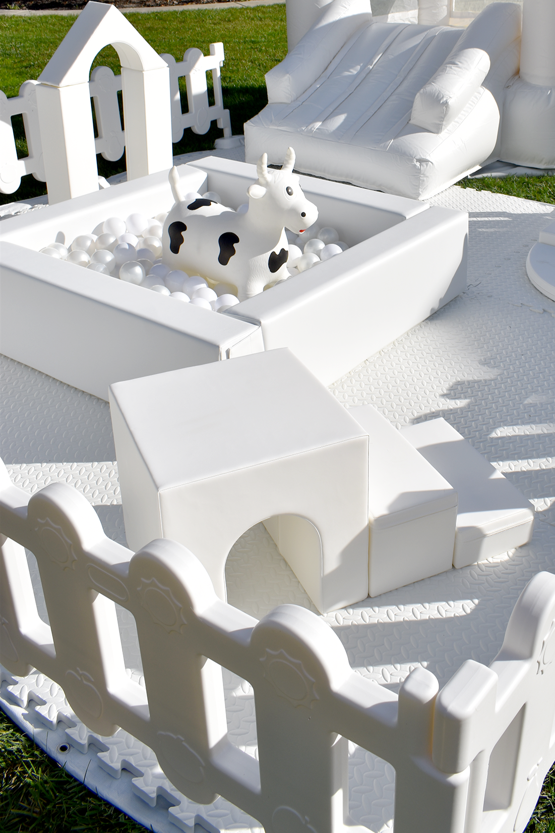White inflatable toy cow in a ball pit, surrounded by white fencing and outdoor play area, with a white inflatable slide and other play structures in the background.