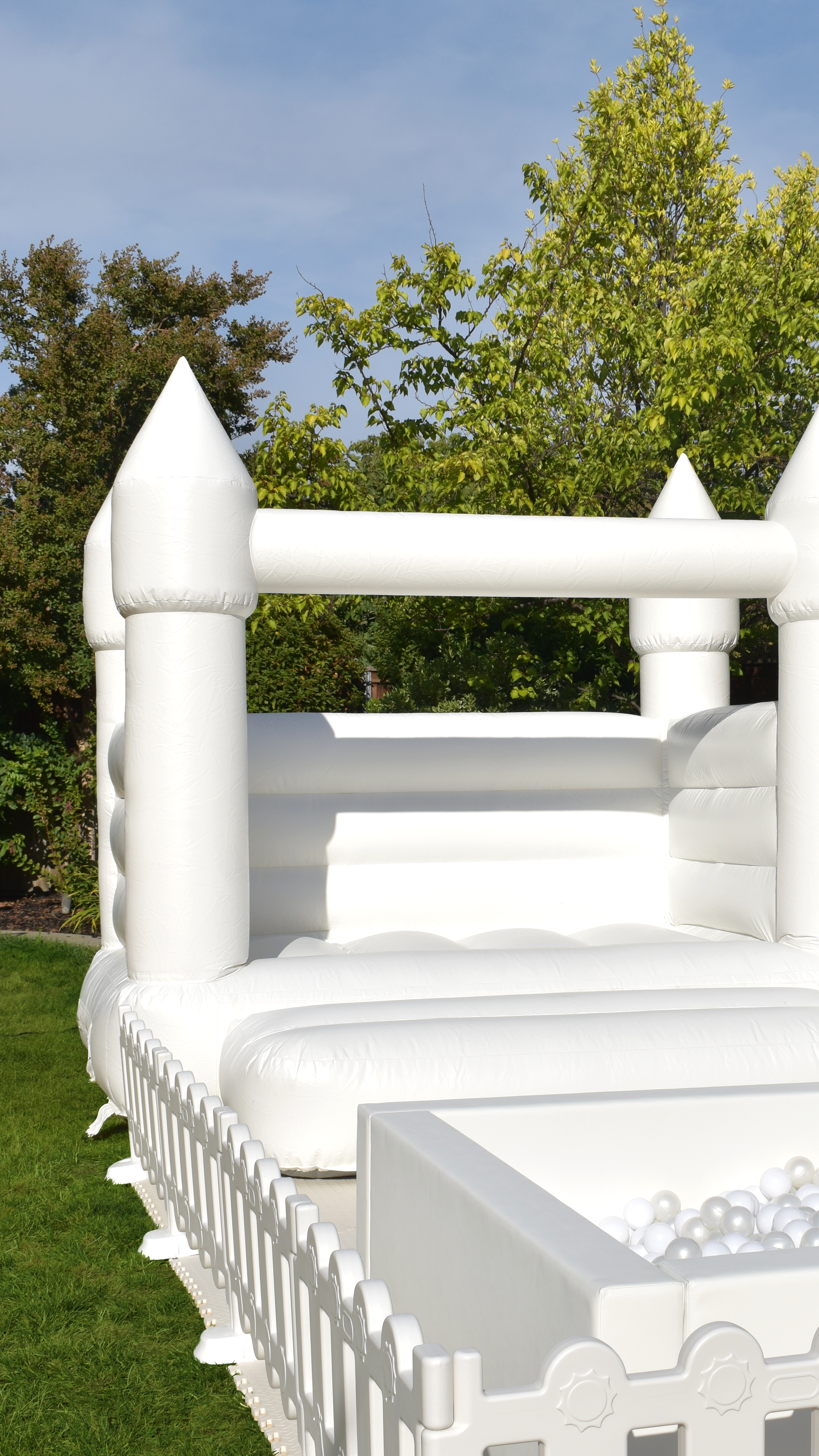 White inflatable castle bounce house with ball pit, set outdoors on grass with trees and blue sky in background.