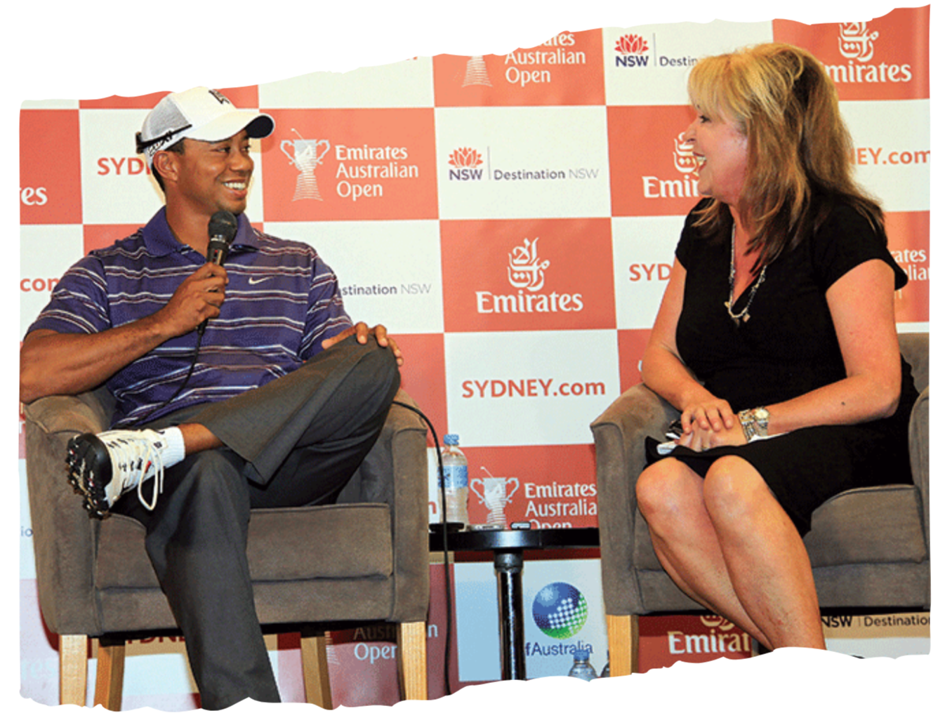 A male golfer and a female interviewer sitting in armchairs during a press interview at the Emirates Australian Open tennis event. The golfer is wearing a striped collared shirt, cap, and golf shoes, holding a microphone, while the interviewer is dressed in a black dress with a necklace and sitting with her hands on her lap. There is a table with a water bottle between them, and a backdrop with logos of the Emirates Australian Open, Sydney, and other sponsors.