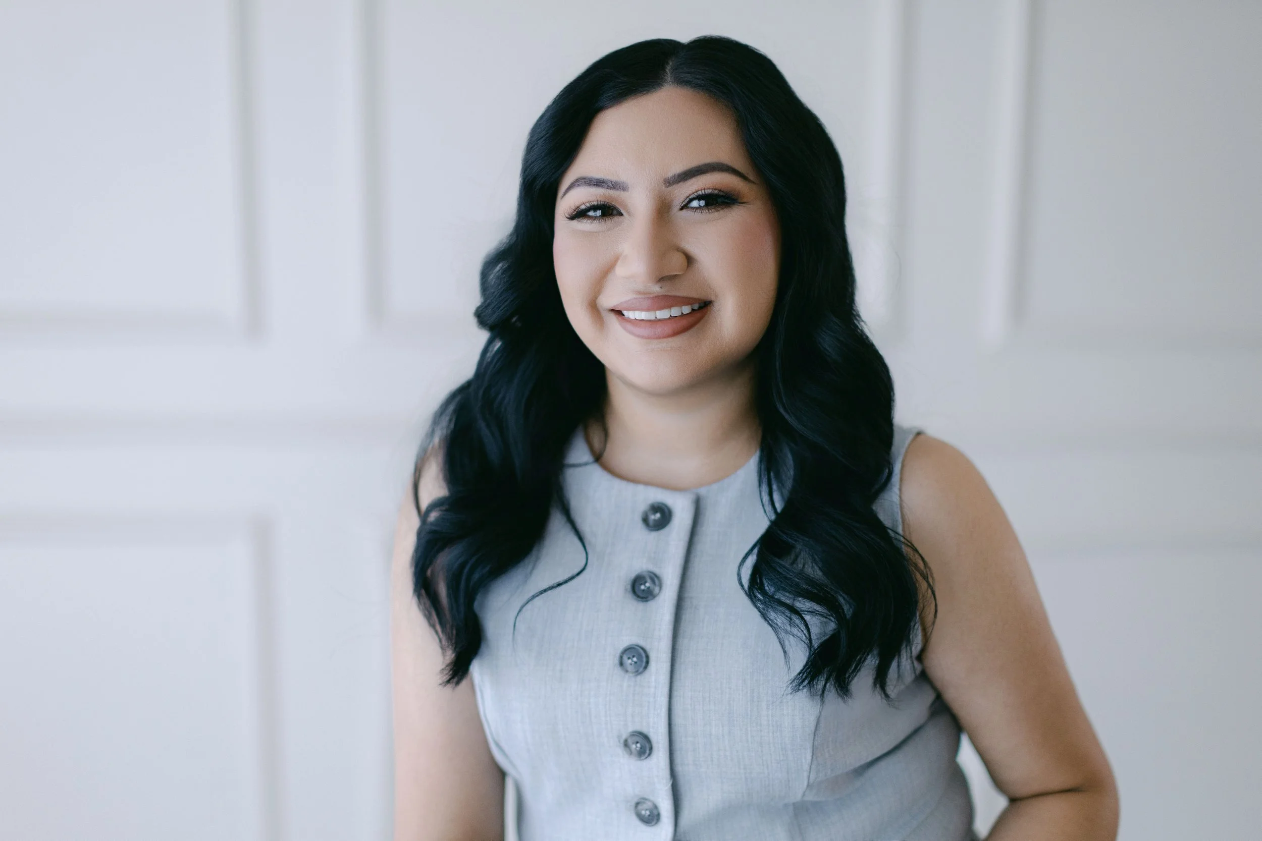A woman with long black wavy hair smiling at the camera, wearing a sleeveless light gray button-up top, standing against a white paneled background.