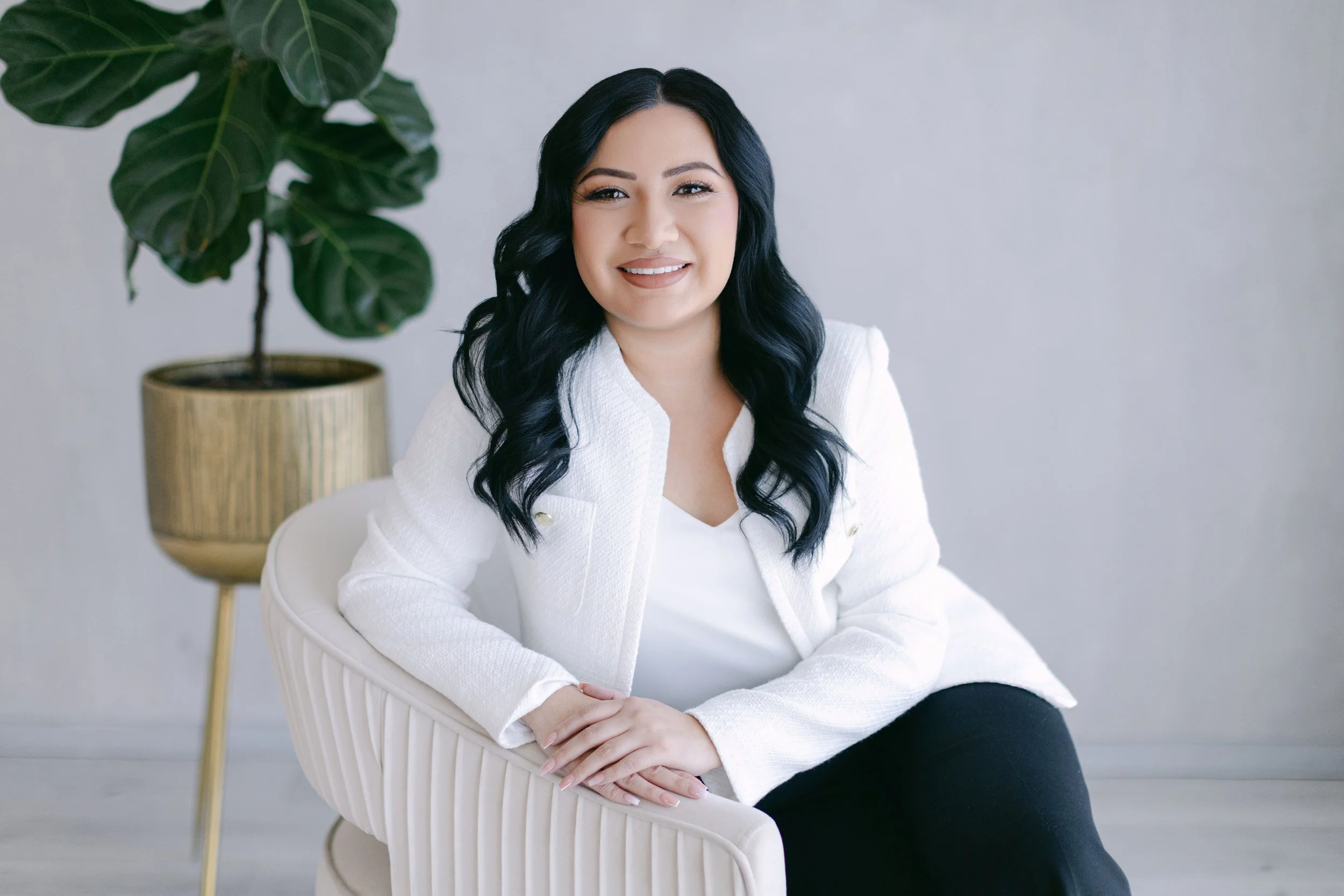 A woman with long black hair, wearing a white blazer, sitting on a light-colored armchair, with a large potted plant in the background.