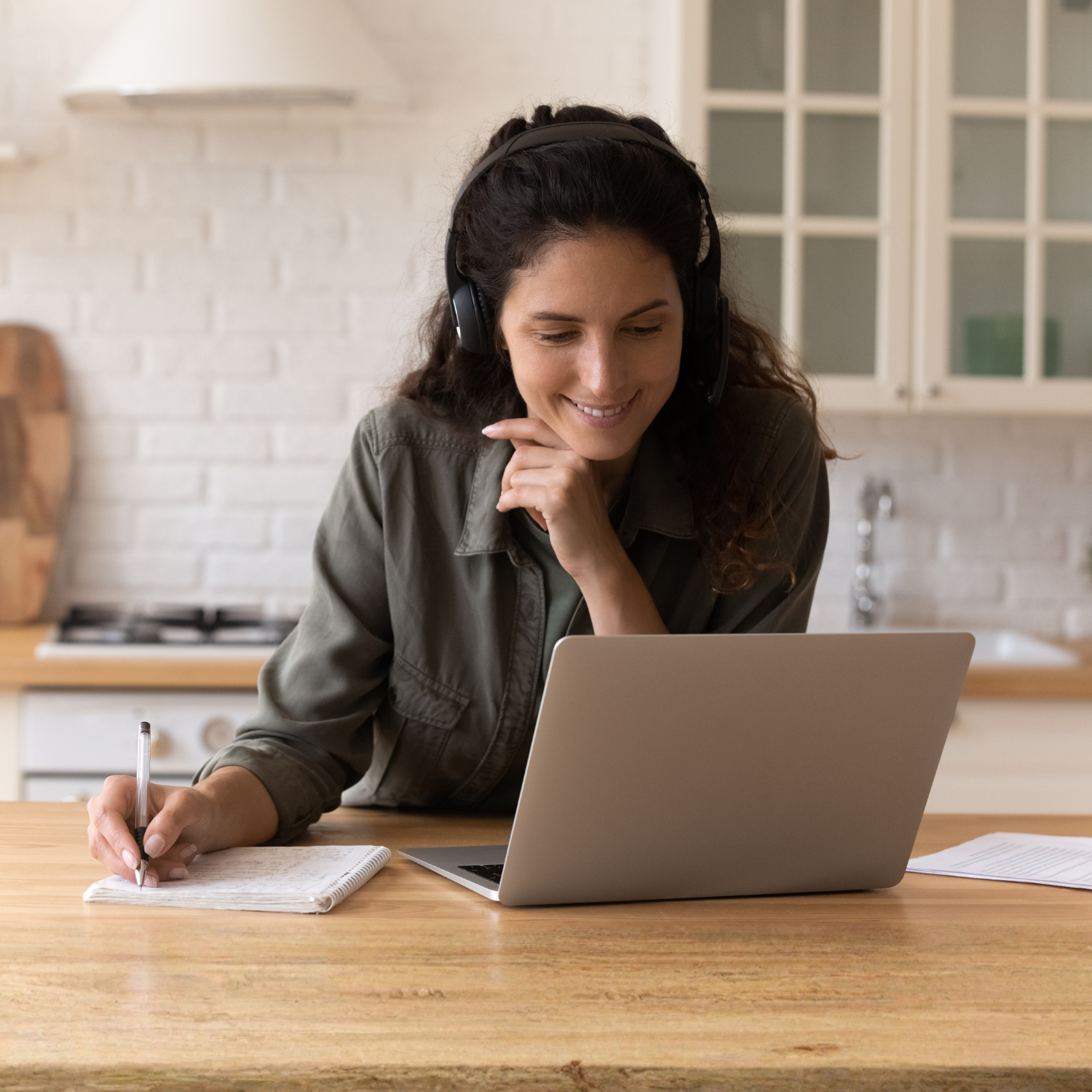 Woman with headphones smiling at her laptop in a kitchen, taking notes on a notepad.