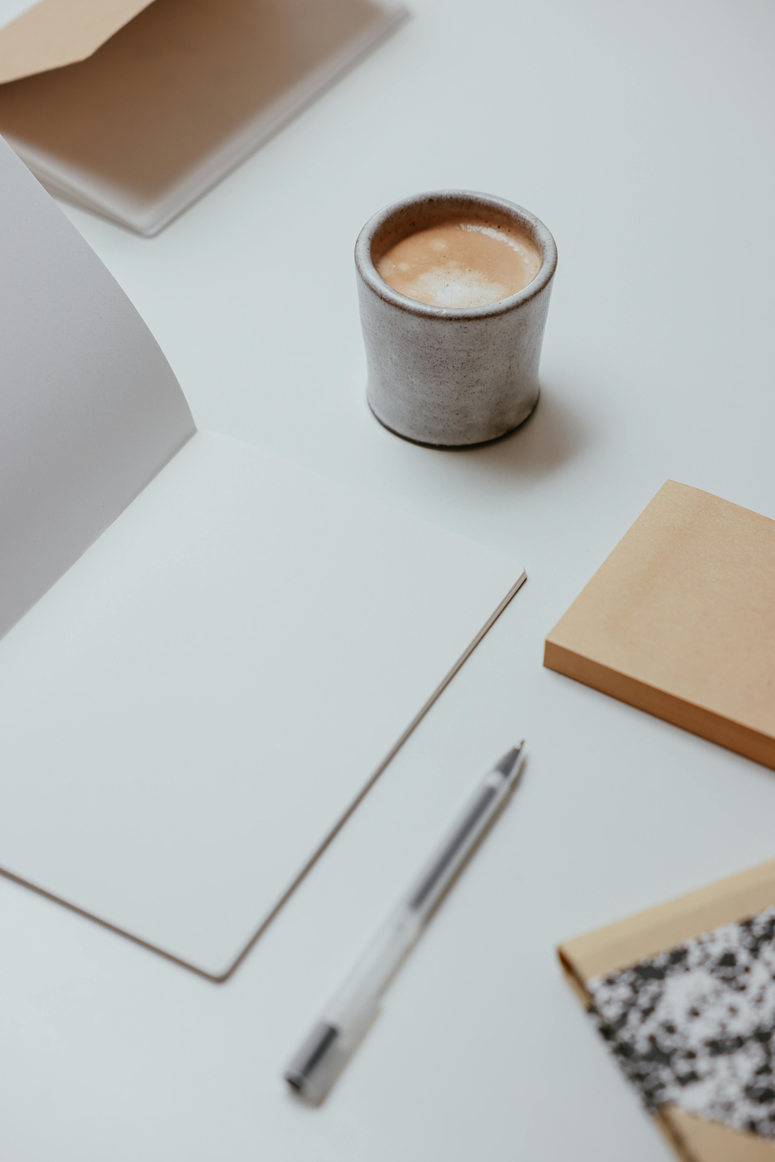 A white desk with a gray ceramic cup filled with coffee, a silver pen, a white notepad, and brown notebooks.