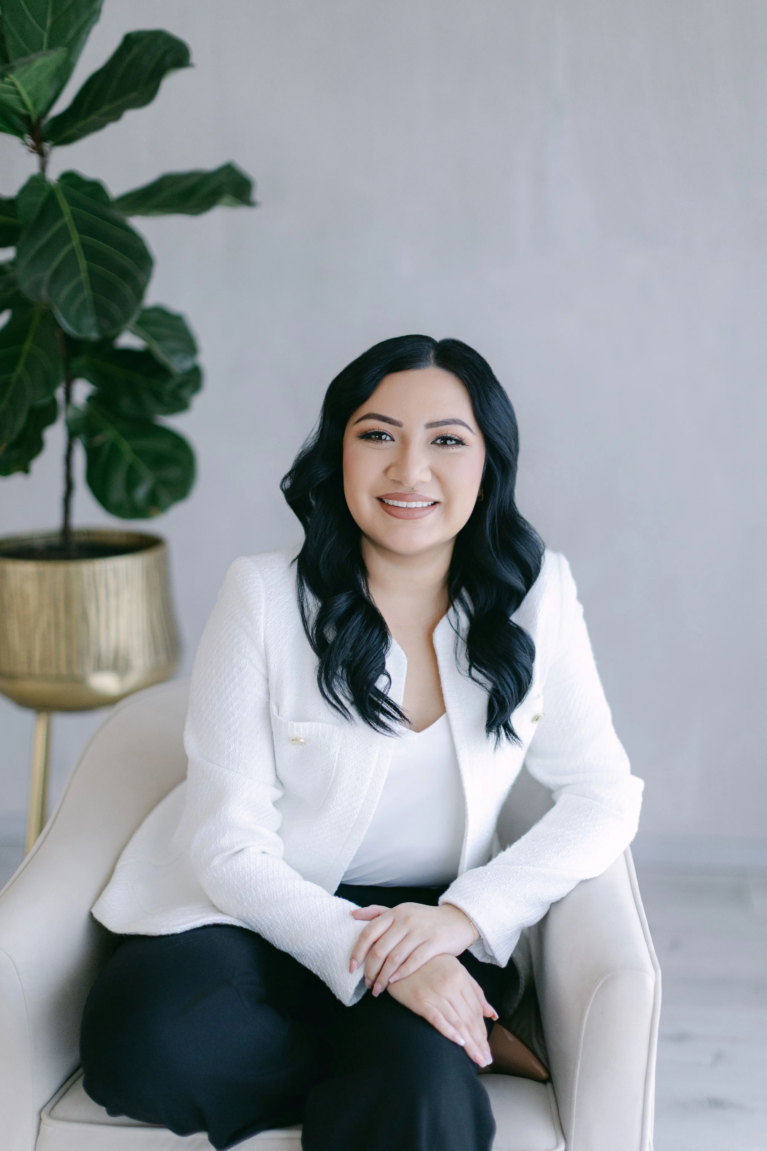 A woman with long black wavy hair, wearing a white blazer and black pants, sitting on a cream-colored armchair, smiling, with a large green plant in a gold pot in the background.