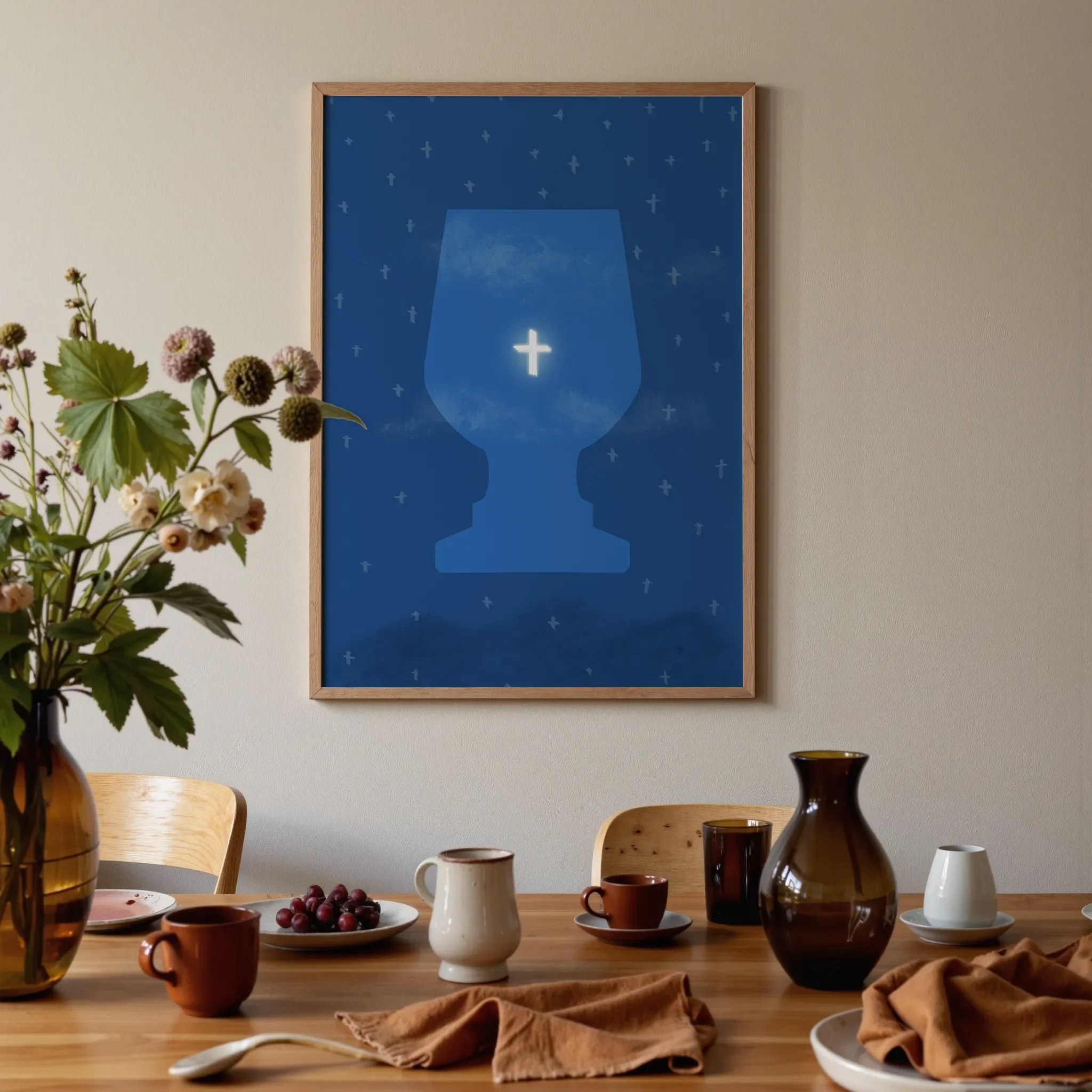 A surreal sky print featuring the cross hangs above a warm wooden dining table with brown and orange tones. On the table are glass vases, ceramics, flowers, and fruit, softly lit by natural light.