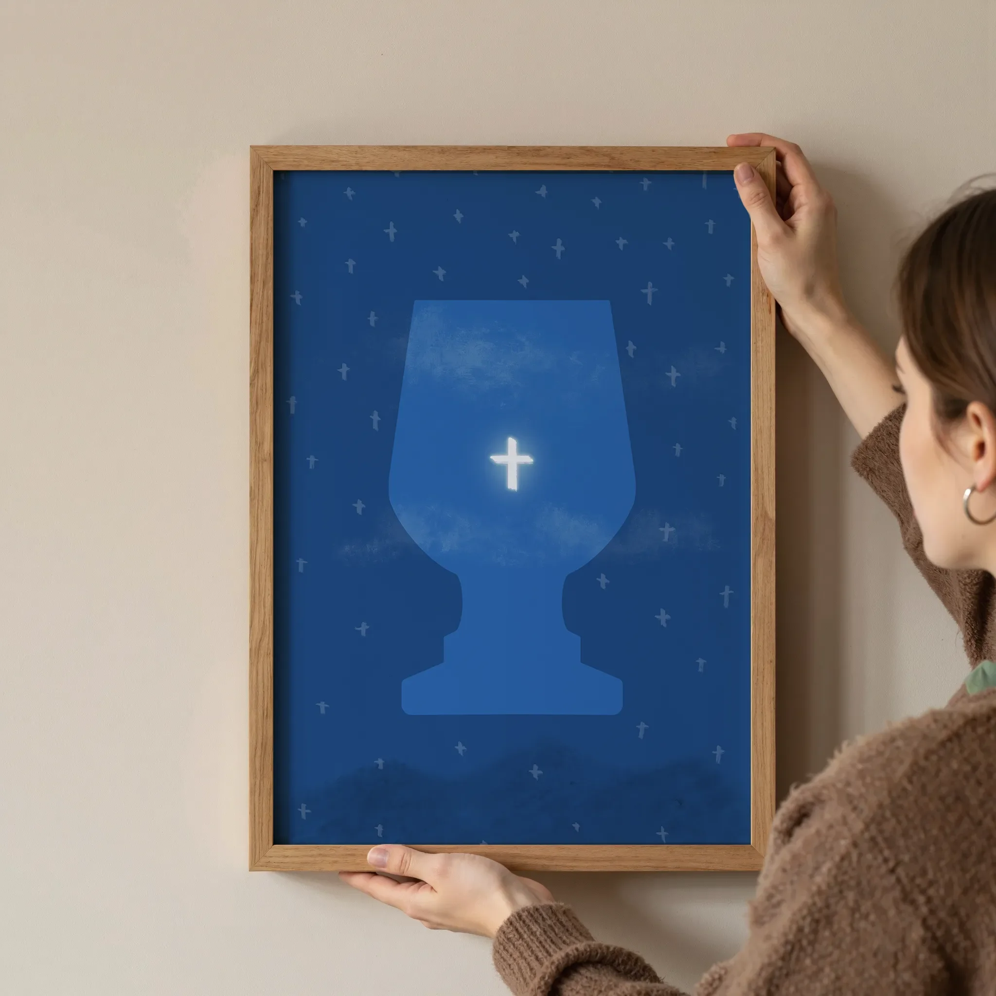 A surreal starry deep blue sky print featuring a bright cross in a translucent cup, shown in a simple wooden frame, is held at the corners by a young woman against a neutral wall.