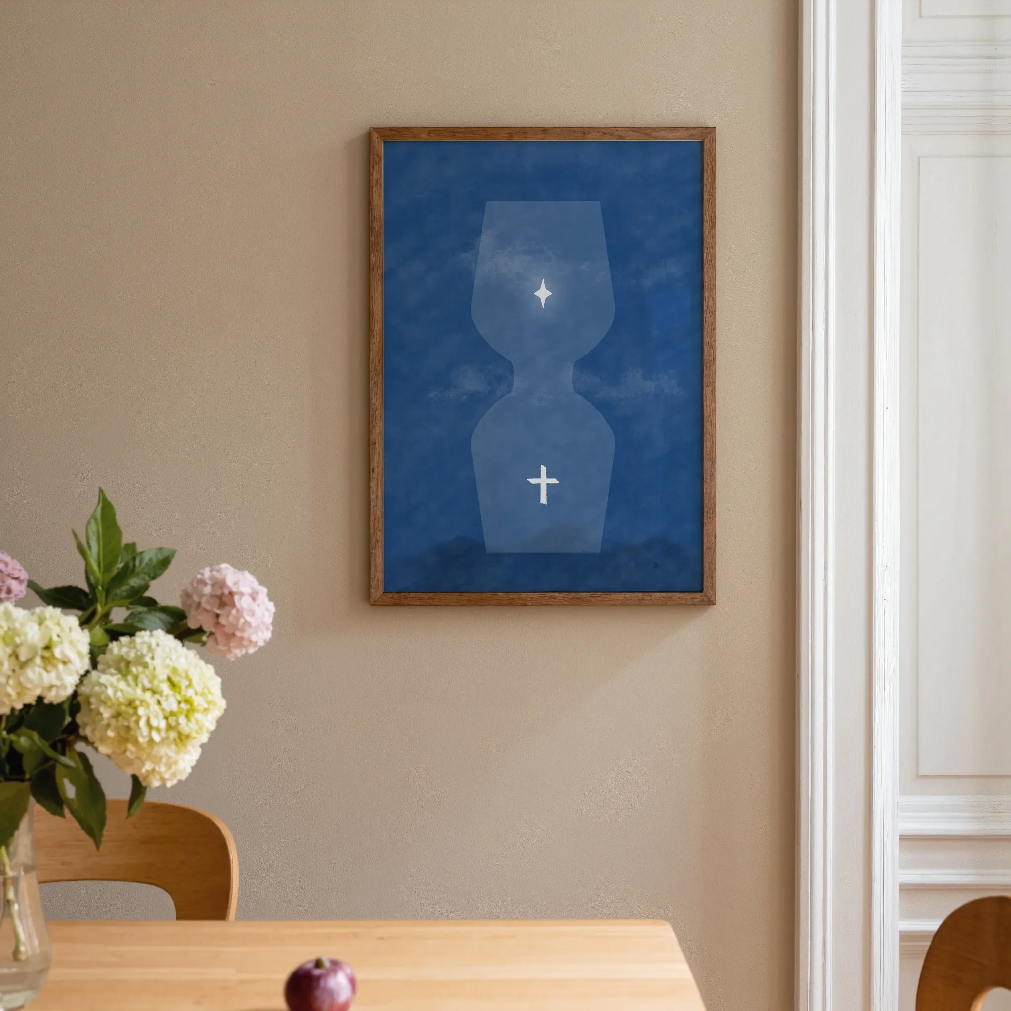 A surreal blue hourglass Christian print hangs on a warm neutral wall beside a white door in a home interior. In front is a warm wooden table with pink and pale yellow flowers. The room is softly lit.