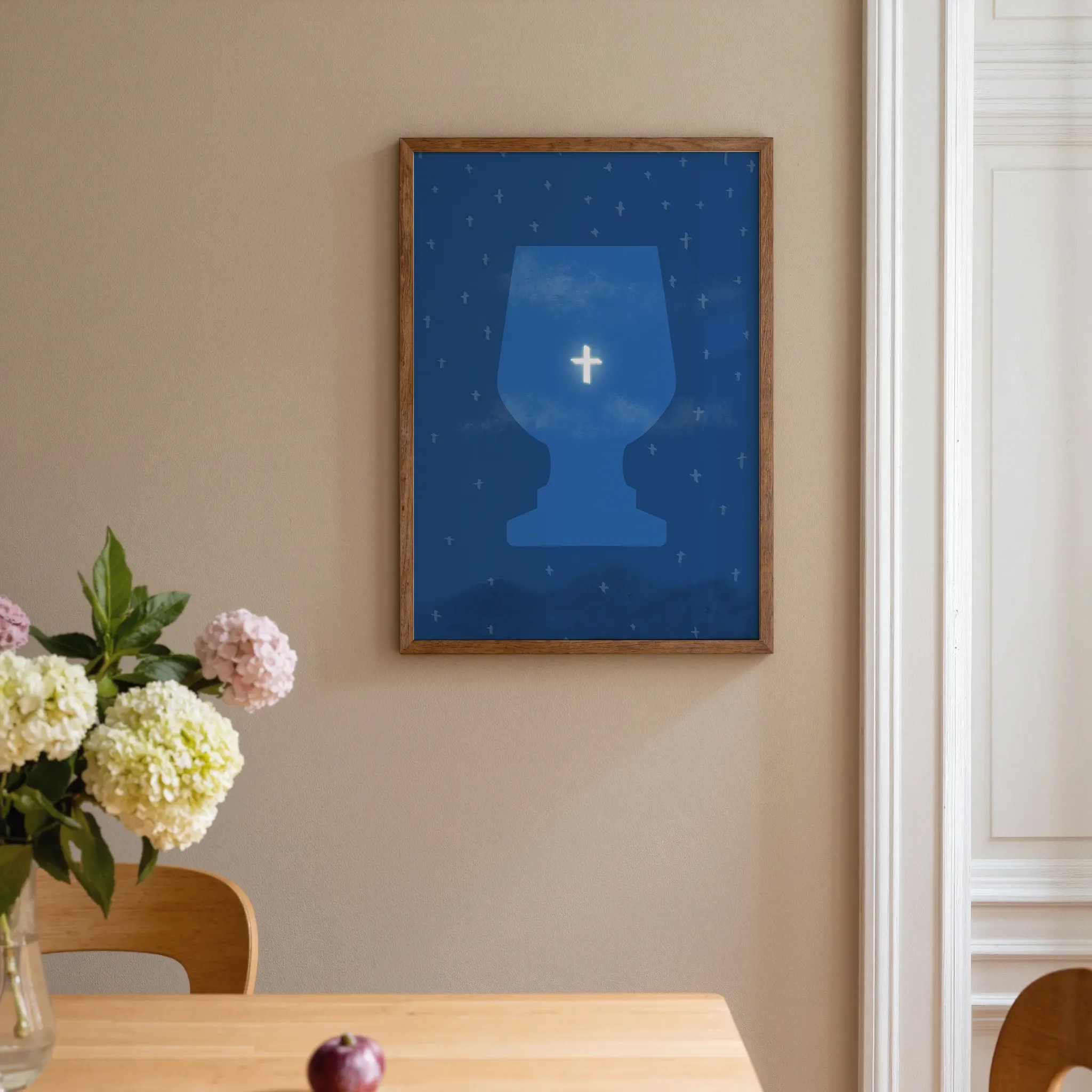 A surreal sky print featuring the cross hangs on a warm neutral wall beside a white door in a home interior. In front is a warm wooden table with pink and pale yellow flowers. The room is softly lit.