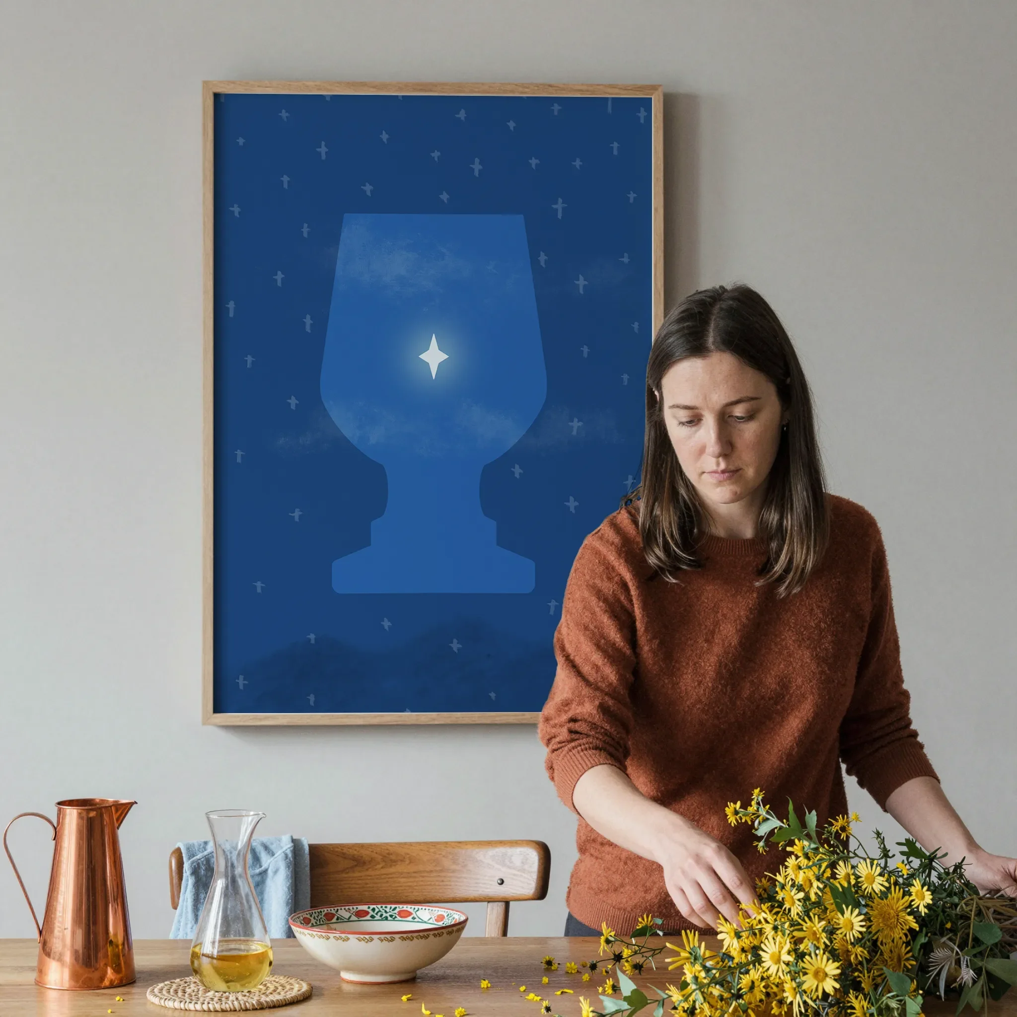 A surreal Star of Bethlehem print hangs on a gray wall in a softly lit dining room. In front, a woman arranges a warm wooden table with yellow flowers, a copper pitcher, ceramic pieces, and glassware.