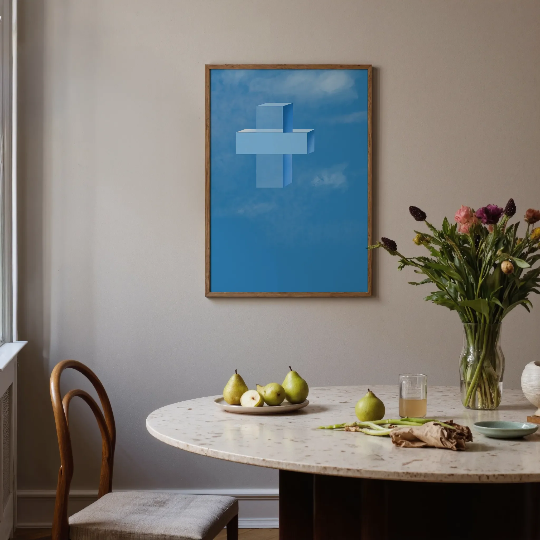 A cross print hangs on a neutral wall in a softly lit room. In front is a round stone table with glassware, ceramics, pears, and flowers. A chair sits on the left.