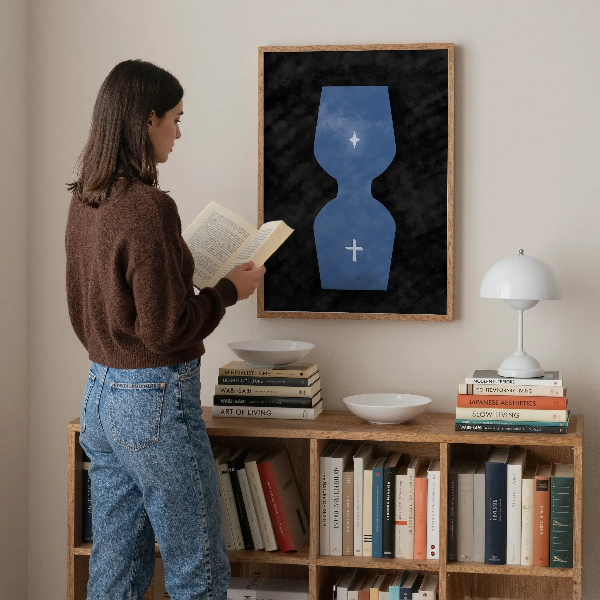 A surreal hourglass Christian print hangs above a wooden bookshelf in a softly lit room. A young woman stands nearby reading a book. On the bookshelf are books, a white lamp, and ceramic bowls.