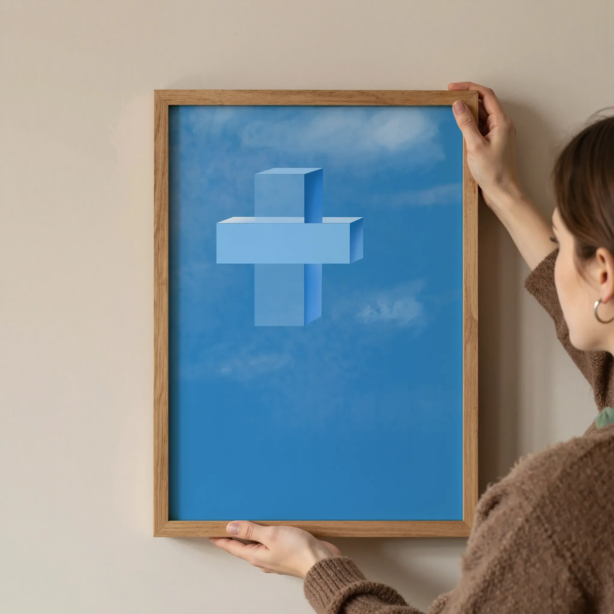 A cross print in a simple wooden frame is held at the corners by a young woman against a neutral wall.