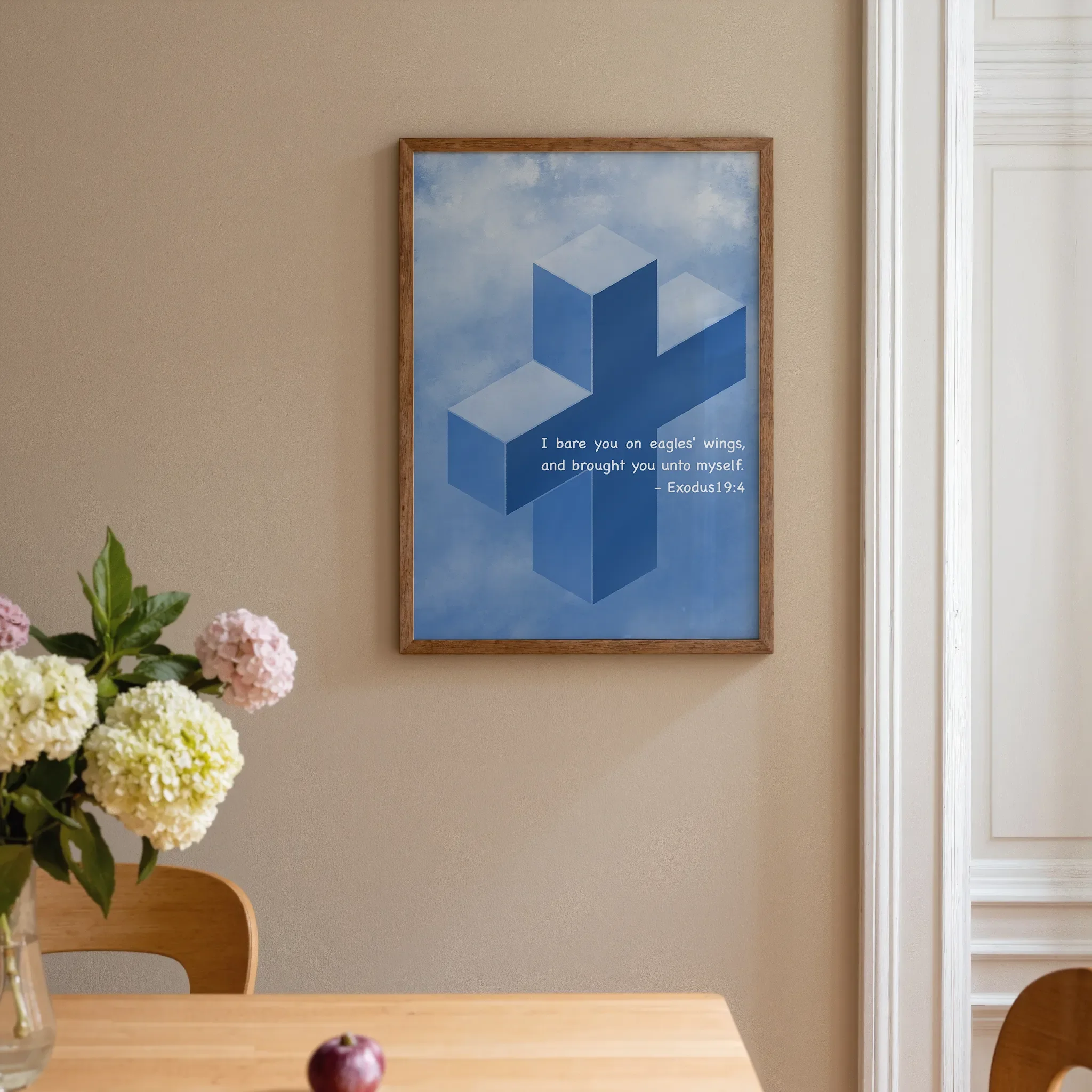 A Scripture wall art featuring a cross hangs on a warm neutral wall beside a white door in a home interior. In front is a warm wooden table with pink and pale yellow flowers. The room is softly lit.