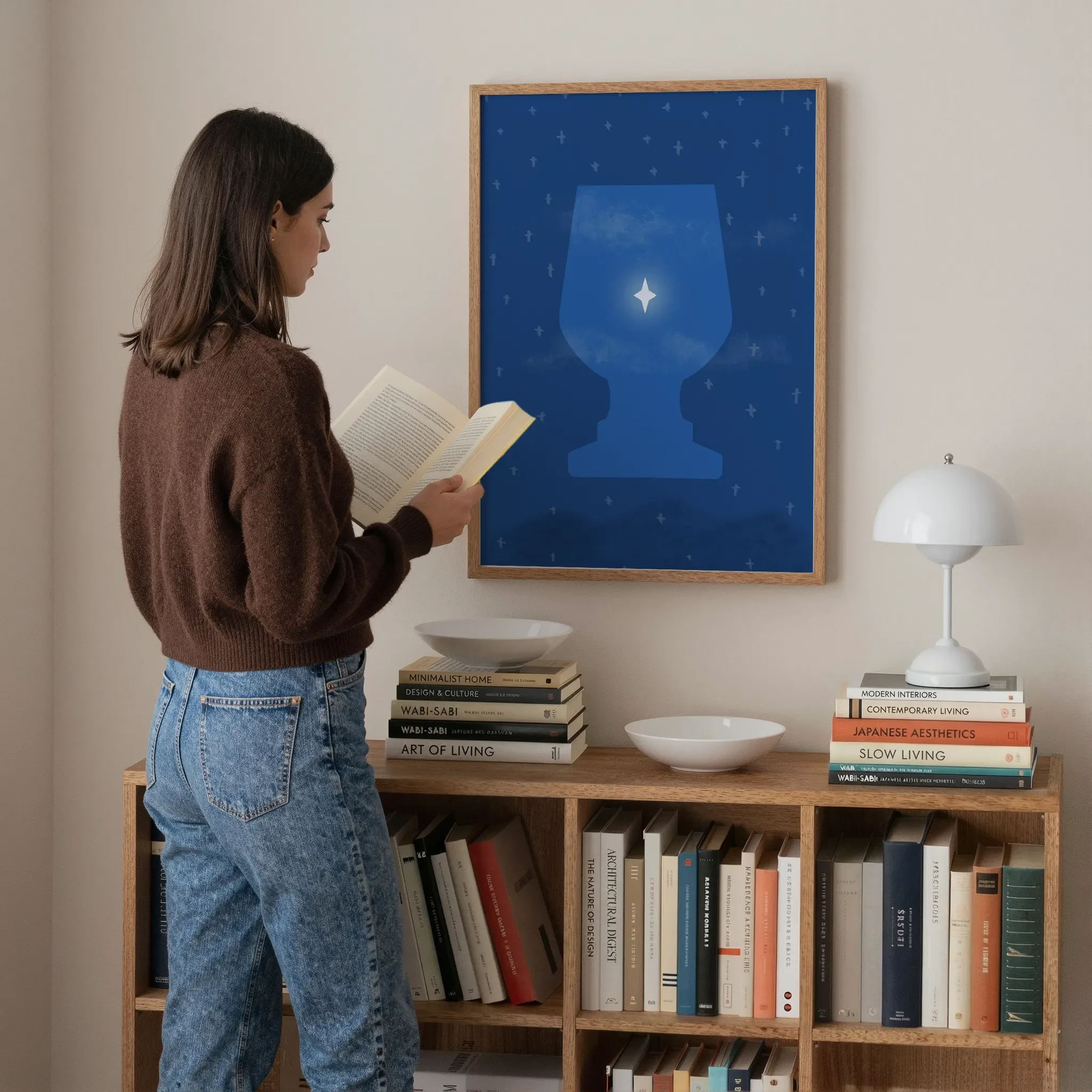 A surreal Star of Bethlehem print hangs above a wooden bookshelf in a softly lit room. A young woman stands nearby reading a book. On the bookshelf are books, a white lamp, and ceramic bowls.