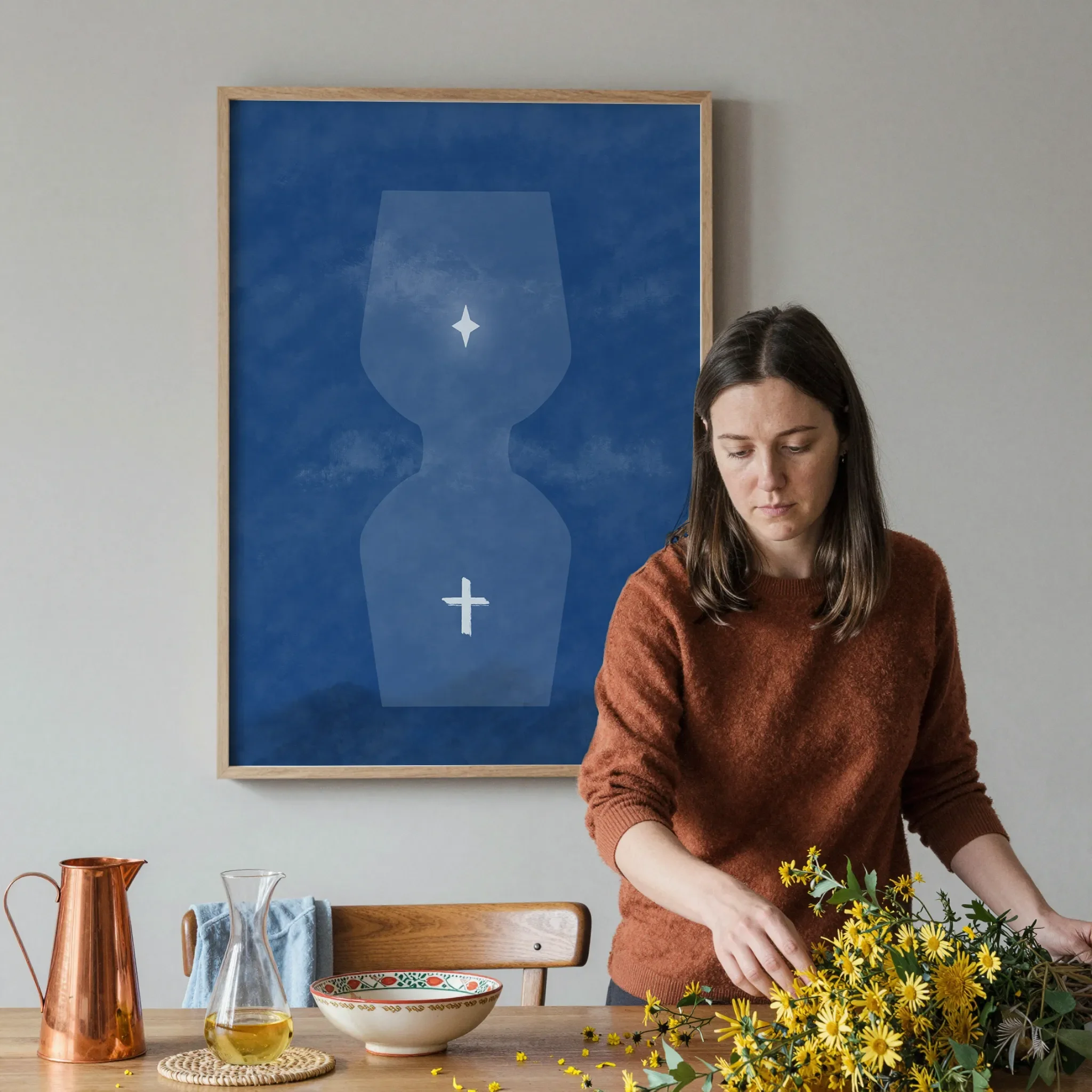 A surreal blue hourglass Christian print hangs on a gray wall in a softly lit dining room. In front, a woman arranges a warm wooden table with yellow flowers, a copper pitcher, ceramics, and glassware