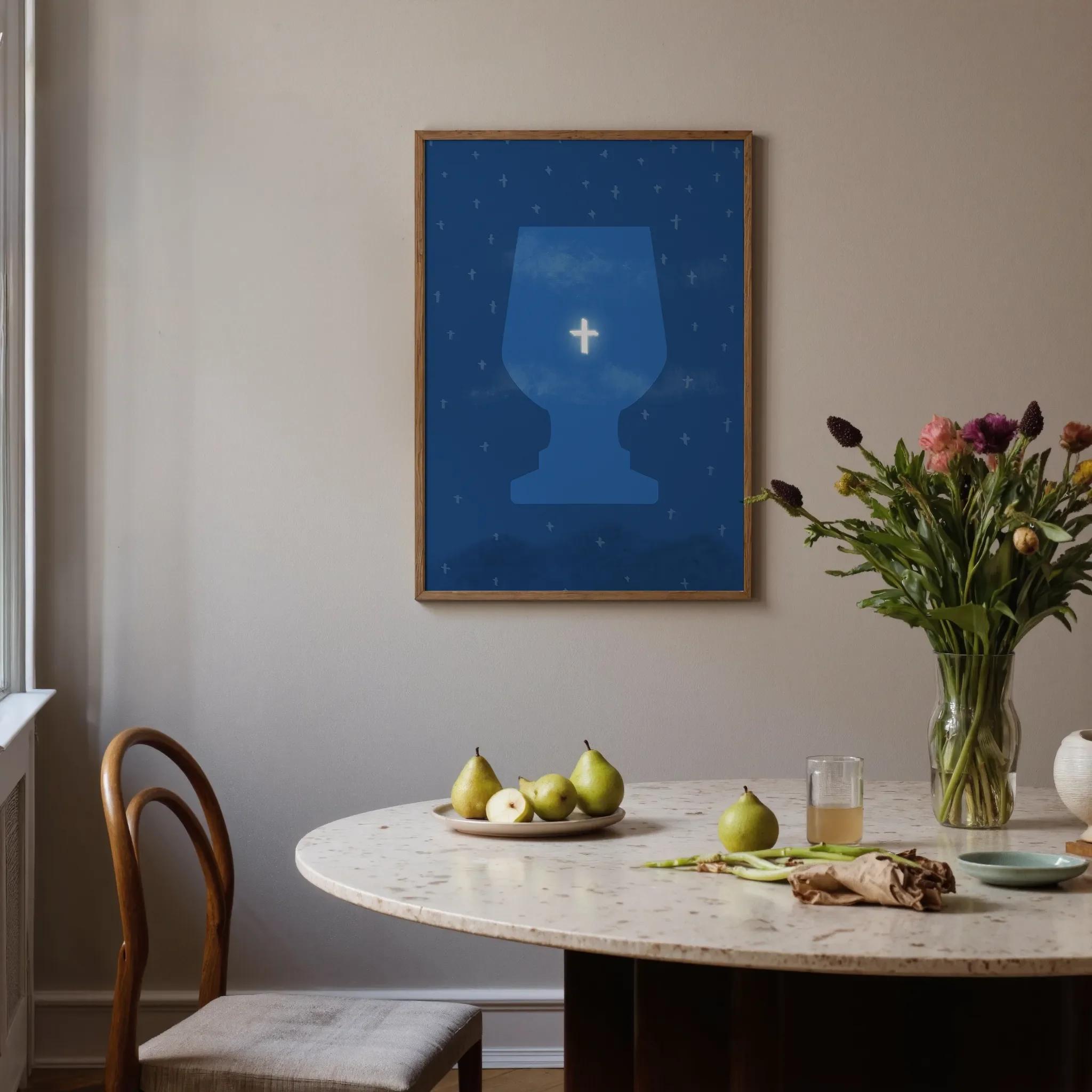 A surreal starry sky print with the cross hangs on a neutral wall in a softly lit room. In front is a round stone table with glassware, ceramic pieces, pears, and flowers. A chair sits on the left.