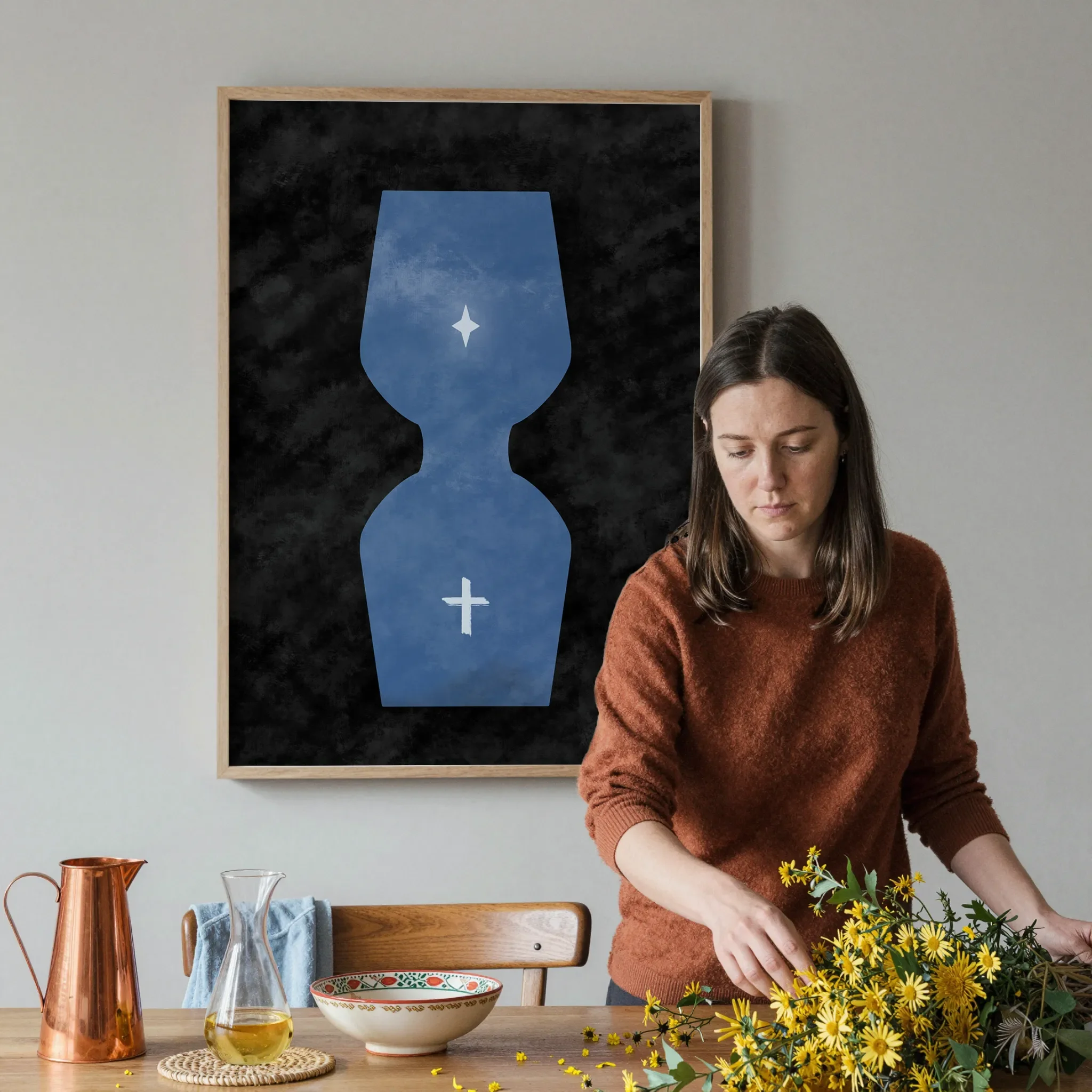 A surreal hourglass Christian print hangs on a gray wall in a softly lit dining room. In front, a woman arranges a warm wooden table with yellow flowers, a copper pitcher, ceramics, and glassware.