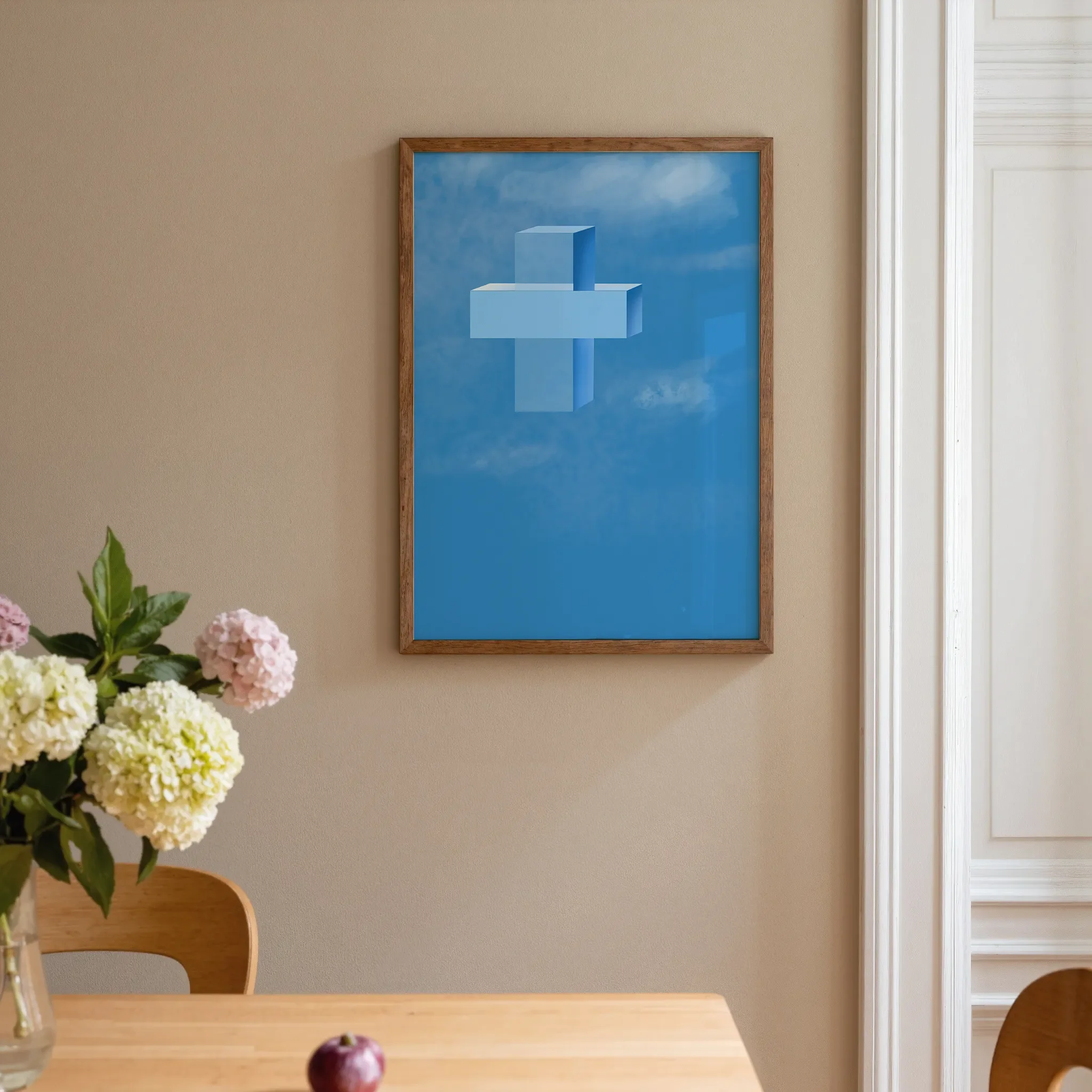 A cross print hangs on a warm neutral wall beside a white door in a home interior. In front is a warm wooden table with pink and pale yellow flowers. The room is softly lit.