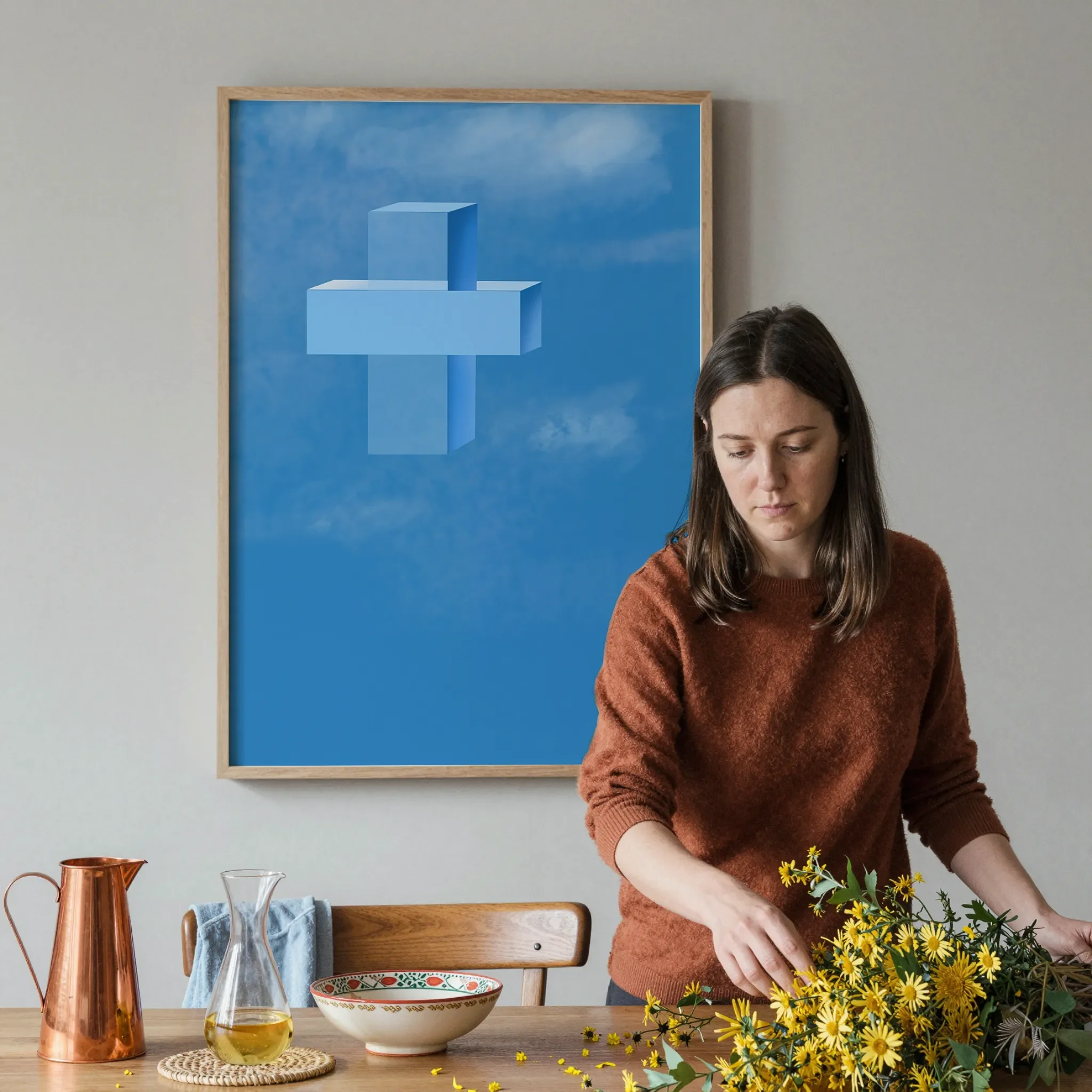 A cross print hangs on a gray wall in a softly lit dining room. In front, a woman arranges a warm wooden table with yellow flowers, a copper pitcher, ceramics, and glassware.