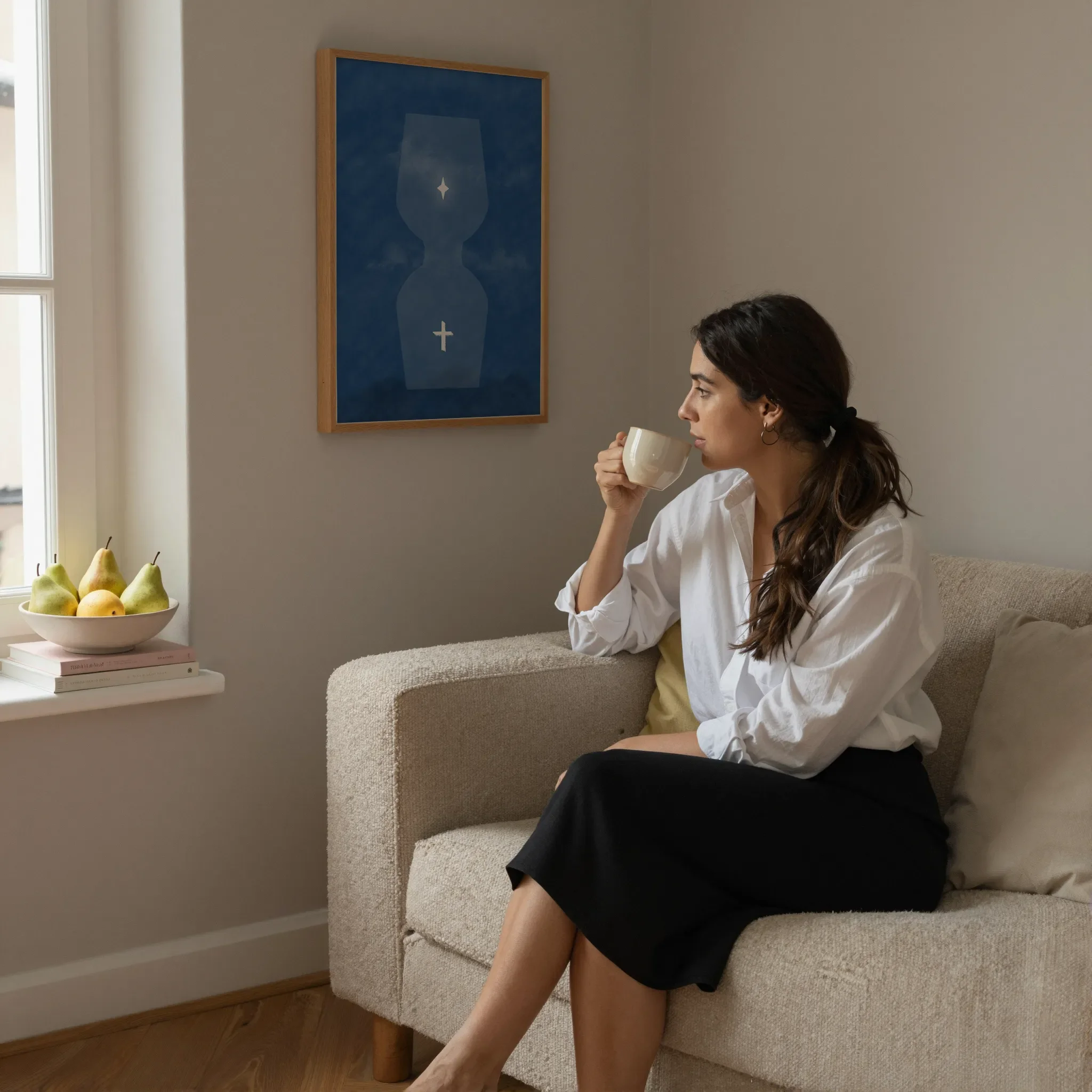 A surreal blue hourglass Christian print hangs on a neutral wall in a cozy living room. A woman sits on a sofa holding a ceramic cup and looking out the window. Pears and books rest on the windowsill.