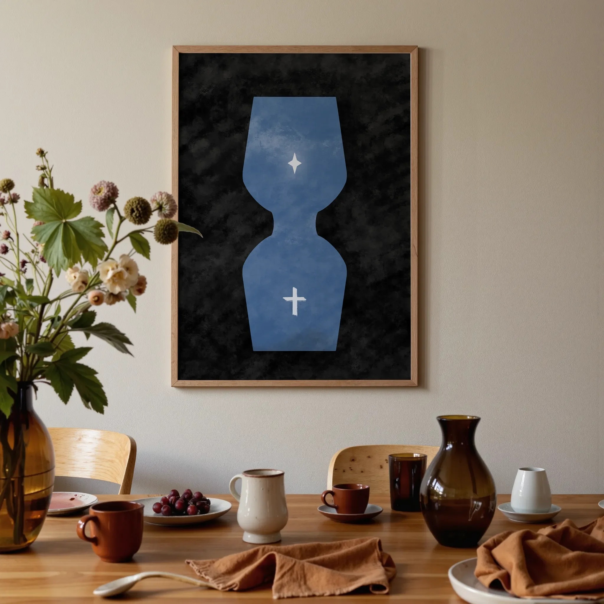 A surreal hourglass Christian print hangs above a warm wooden dining table in brown and orange tones. On the table are glass vases, ceramic pieces, flowers, and fruit, softly lit by natural light.