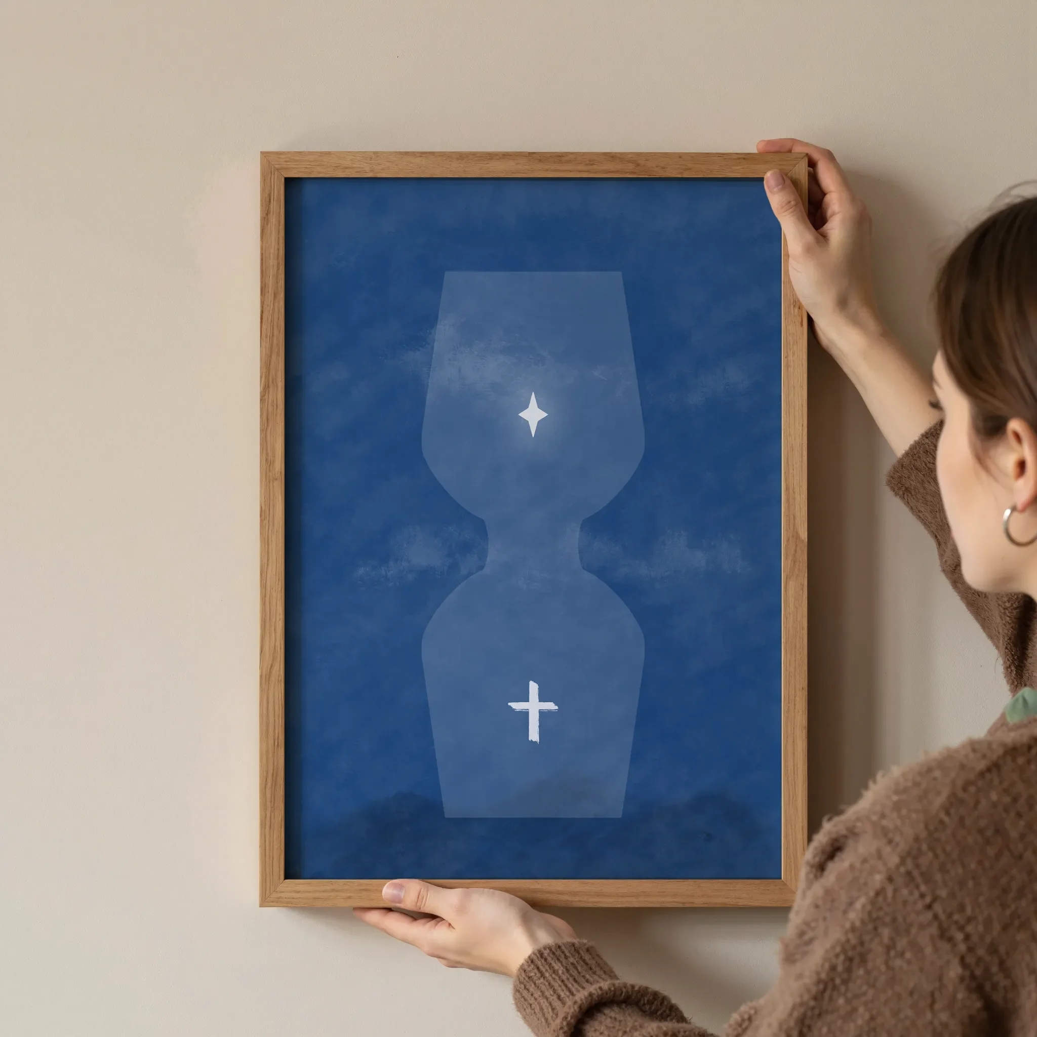 A surreal blue hourglass Christian print with a star above and a glowing cross below, shown in a simple wooden frame, is held at the corners by a young woman against a neutral wall.